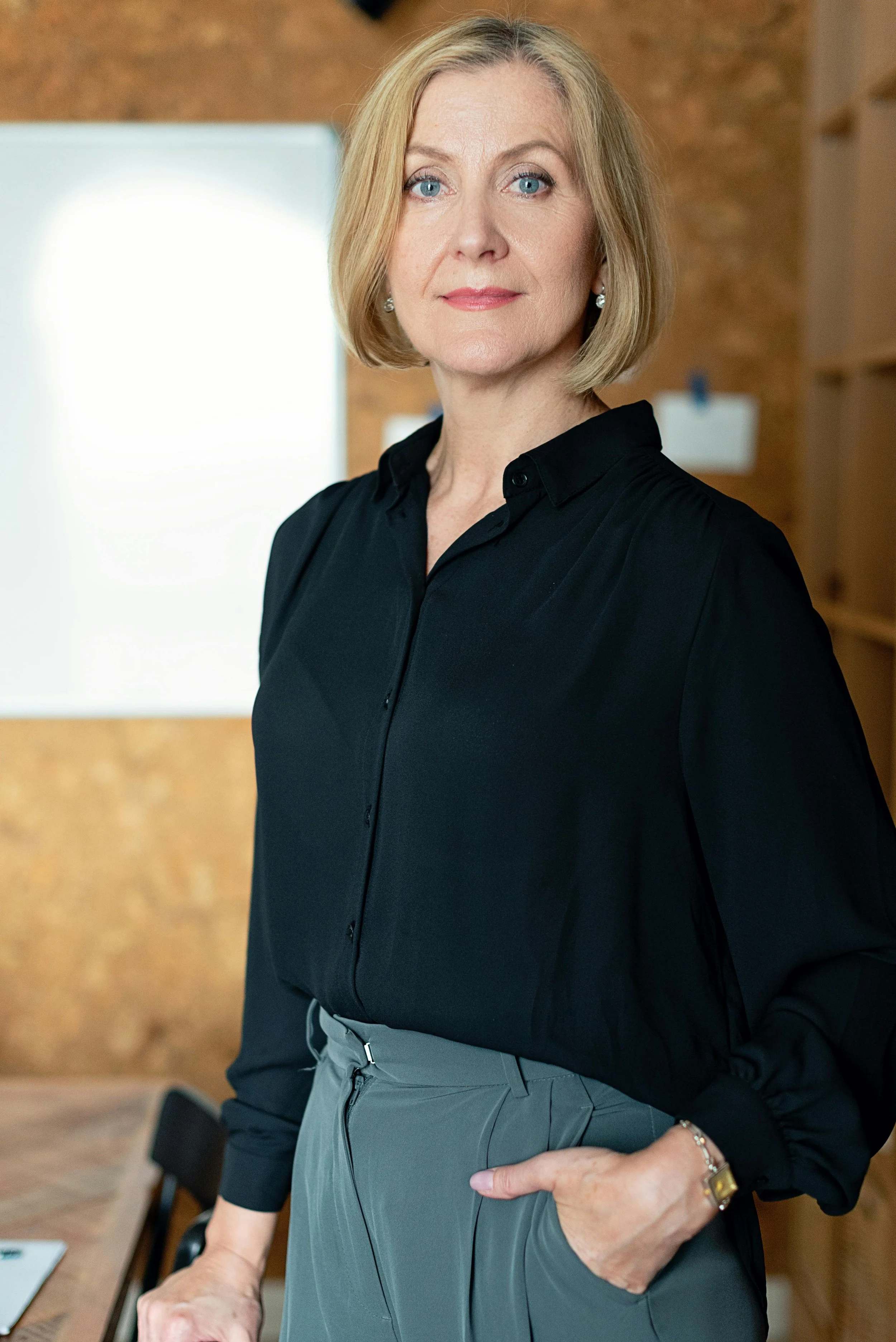 A middle-aged woman with blonde hair and blue eyes wearing a black blouse and gray pants, standing in a room with wooden walls.