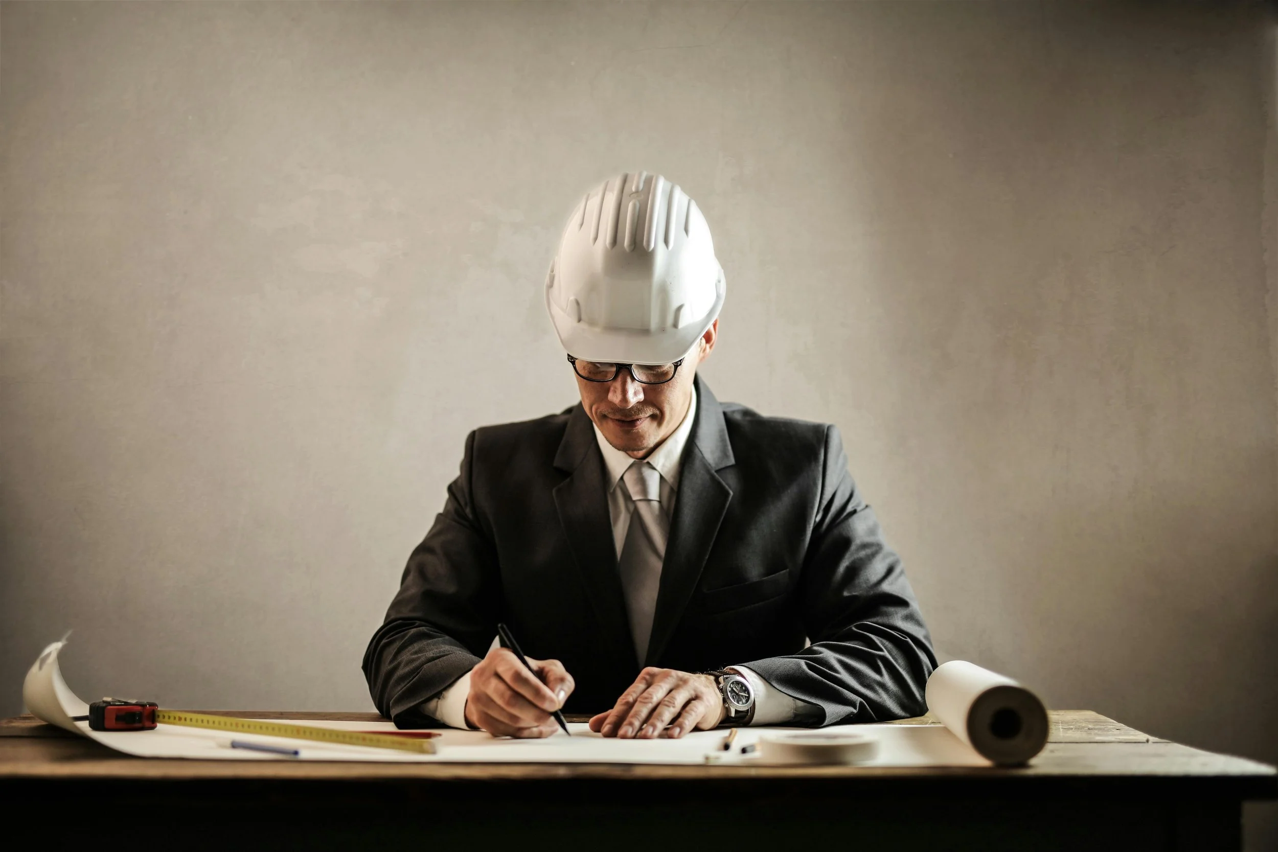 A man in a suit and tie, wearing a white safety helmet and glasses, is sitting at a desk, drawing or writing on a large sheet of paper with various drafting tools and blueprints on the desk.
