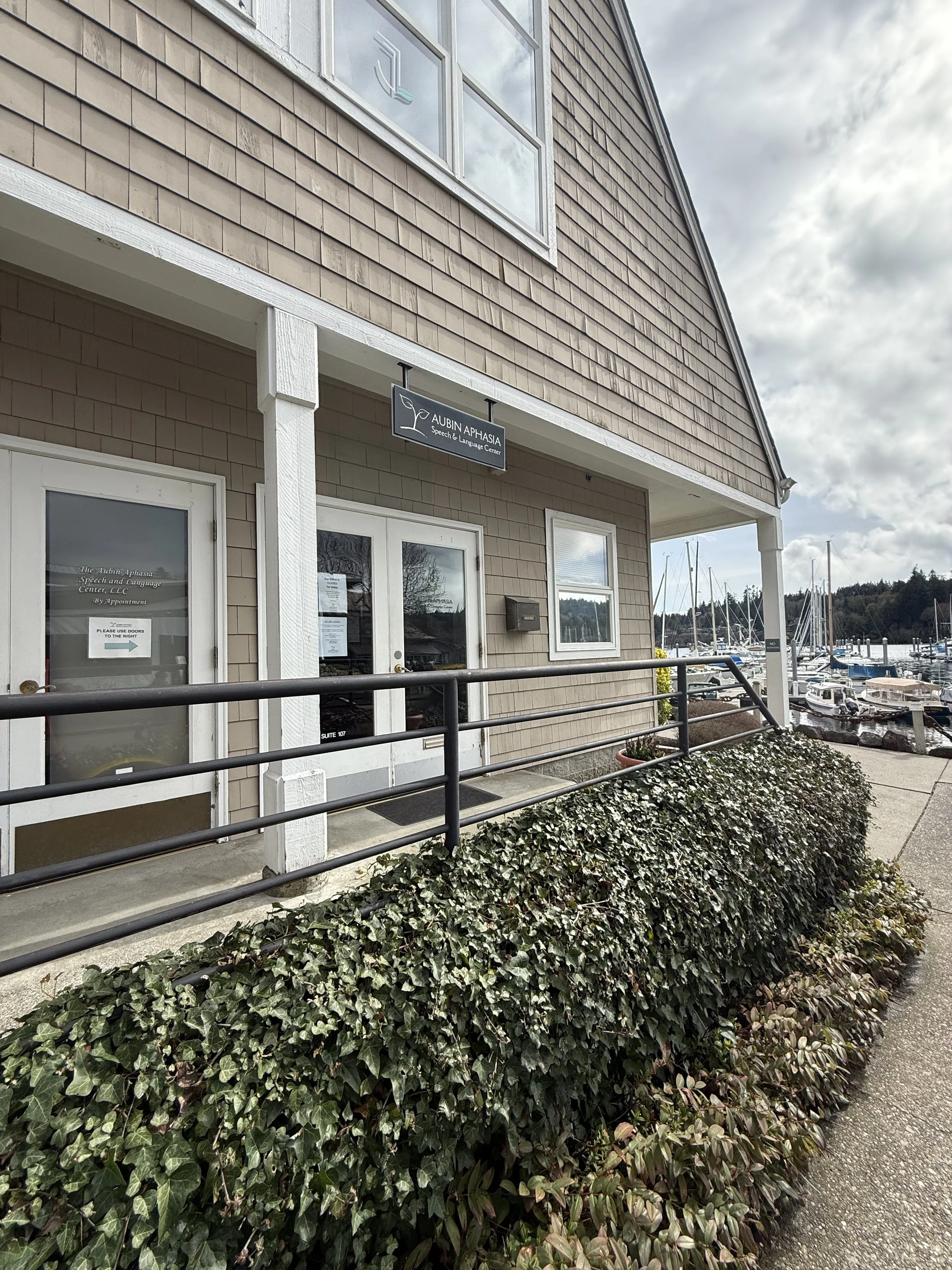 The exterior of a speech and language center building with a sign that reads 'Aubin Aphasia Speech & Language Center LLC by Appointment'. The building has beige siding and white trim, with glass double doors and a window above. There is a pathway with a metal railing, bushes, and a marina with boats in the background under a cloudy sky.