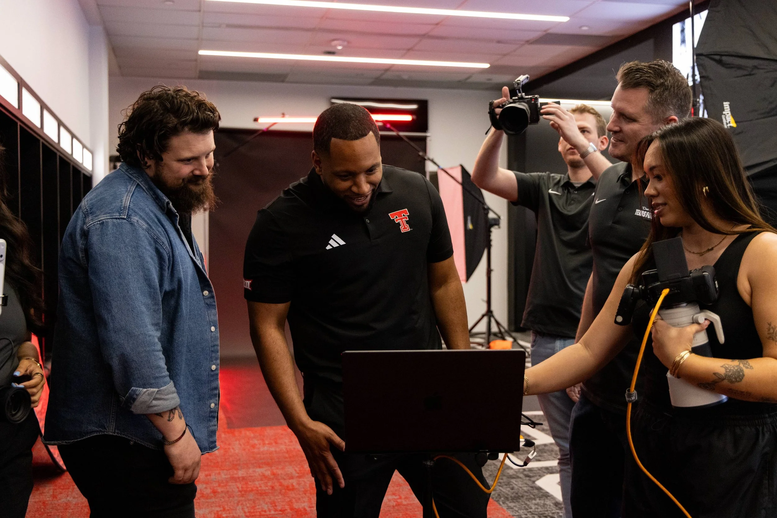 A group of people, including a man in black shirt with Texas Tech logo, gathered around a laptop in an indoor setting with professional lighting and photography equipment.
