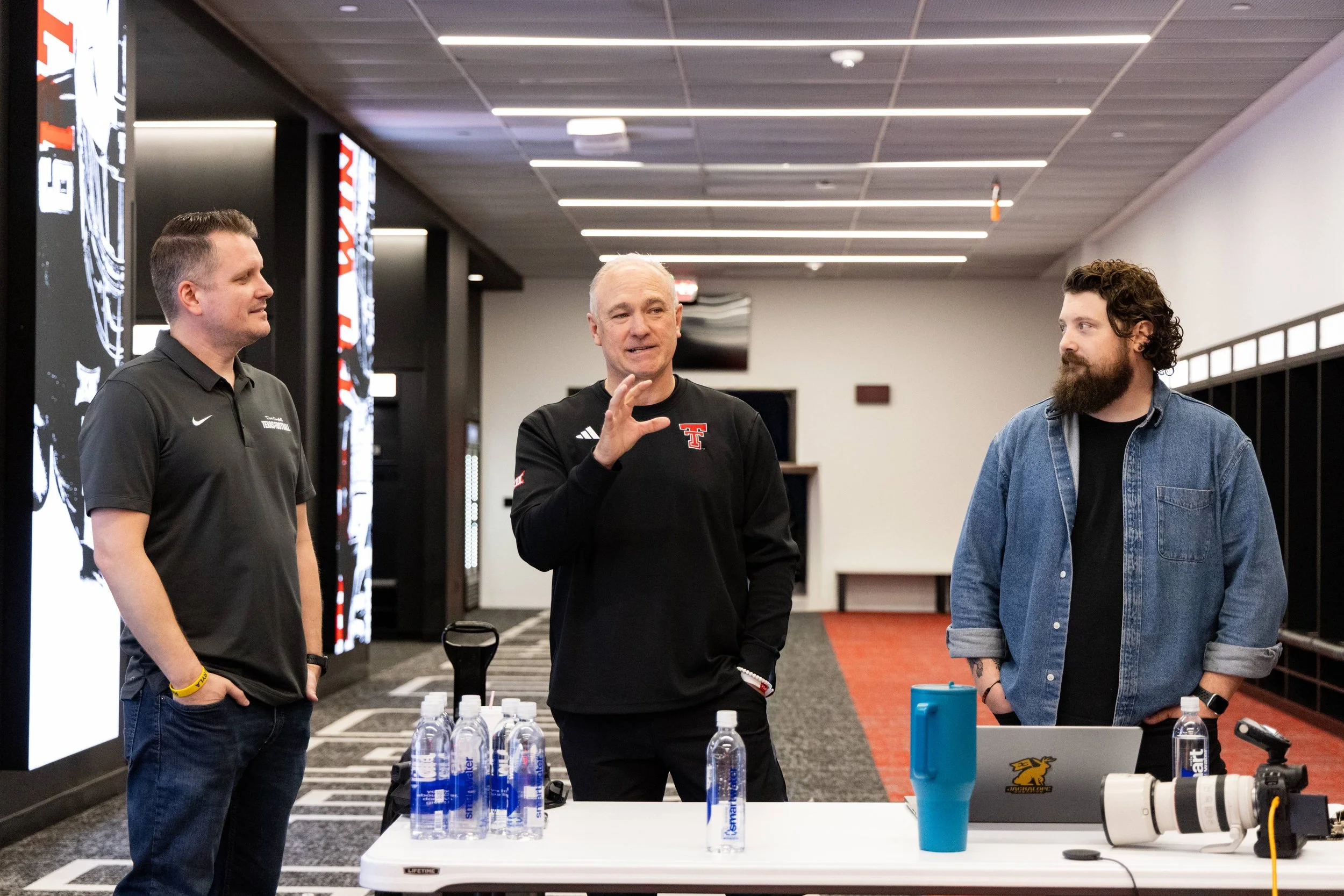 Three men are standing in a room with a table that has several water bottles on it. The man in the middle is talking, while the other two listen. The room has digital displays and a ceiling with linear lighting fixtures.