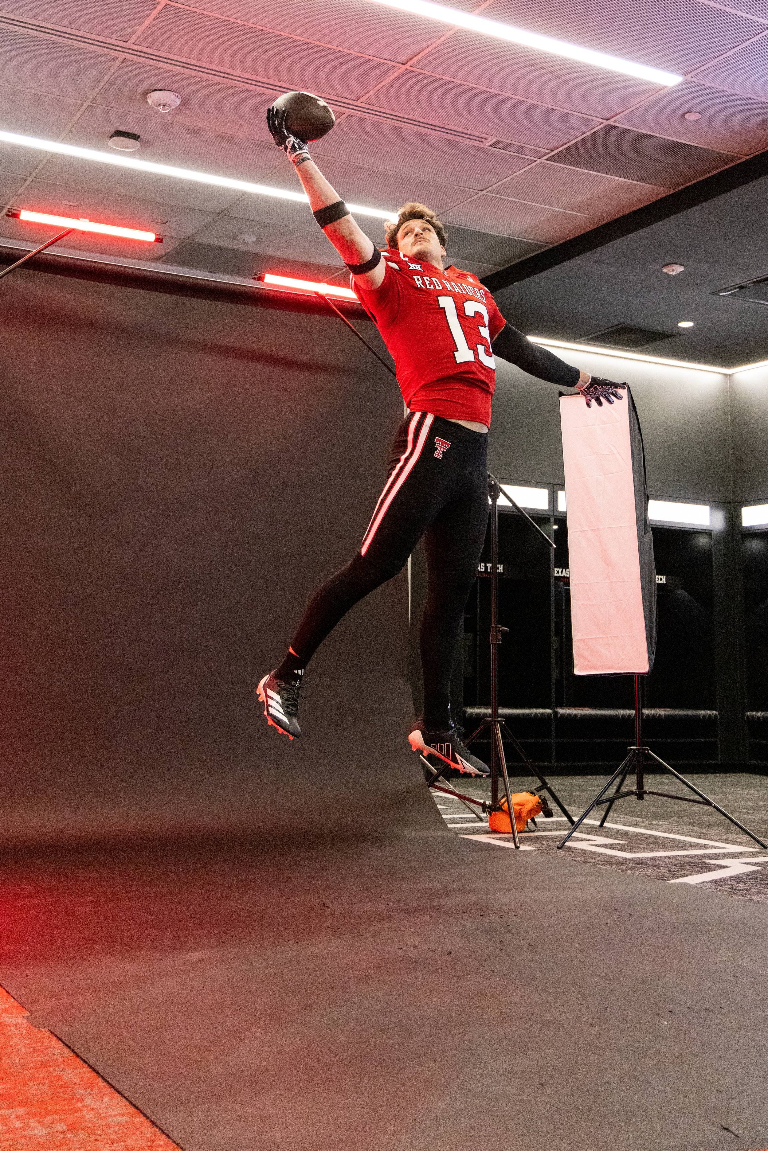Football player in a red uniform with the number 13 and 'Red Raiders' on it, jumping for a catch during a photoshoot in a studio with lighting equipment and black walls.