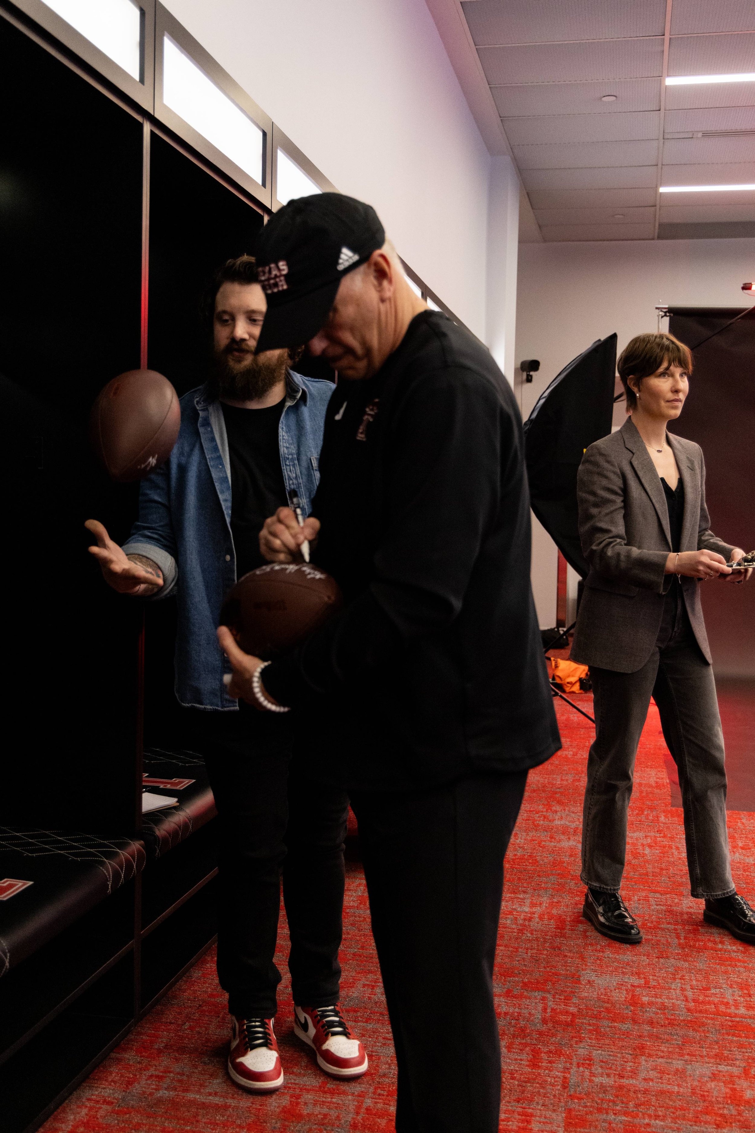 Three people in a room with a red carpet, two men and one woman. One man, wearing a black cap and black jacket, is signing a football while the other, with a beard and denim jacket, looks on. The woman, dressed in a grey blazer and black pants, stand