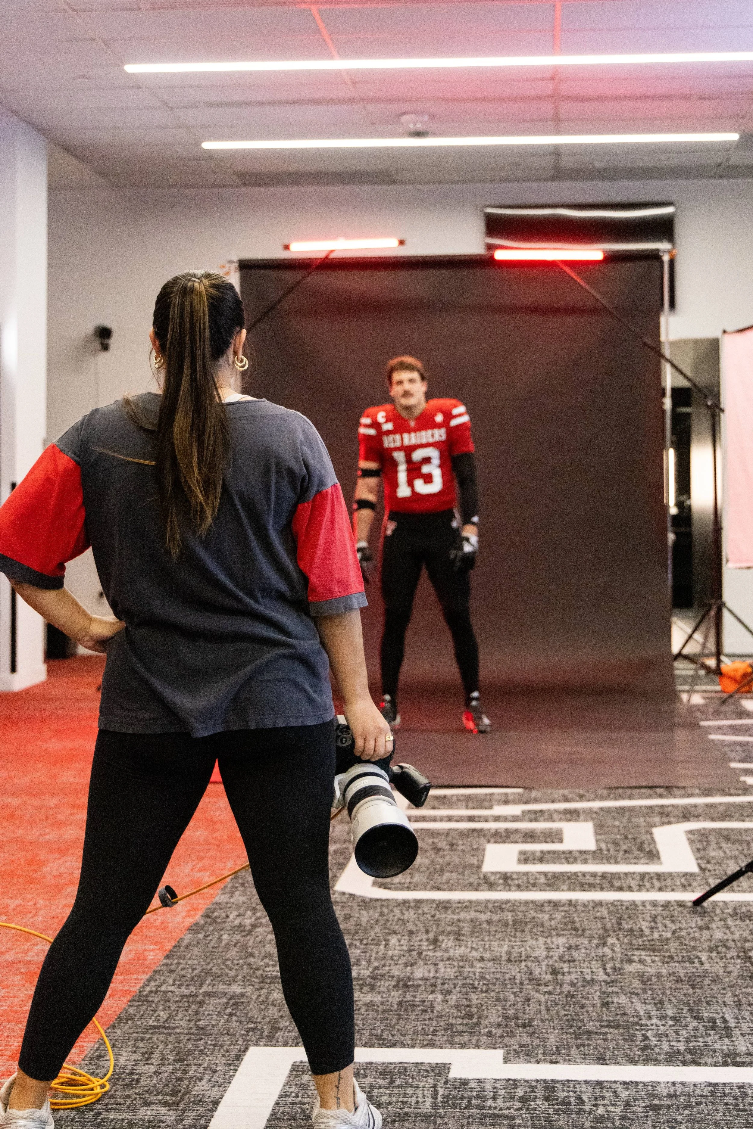 A woman with long dark hair in a ponytail, wearing black pants, a gray and red sports shirt, and sneakers, is holding a camera with a large telephoto lens. She is standing on a gray and red patterned floor, facing a football player in a red jersey wi