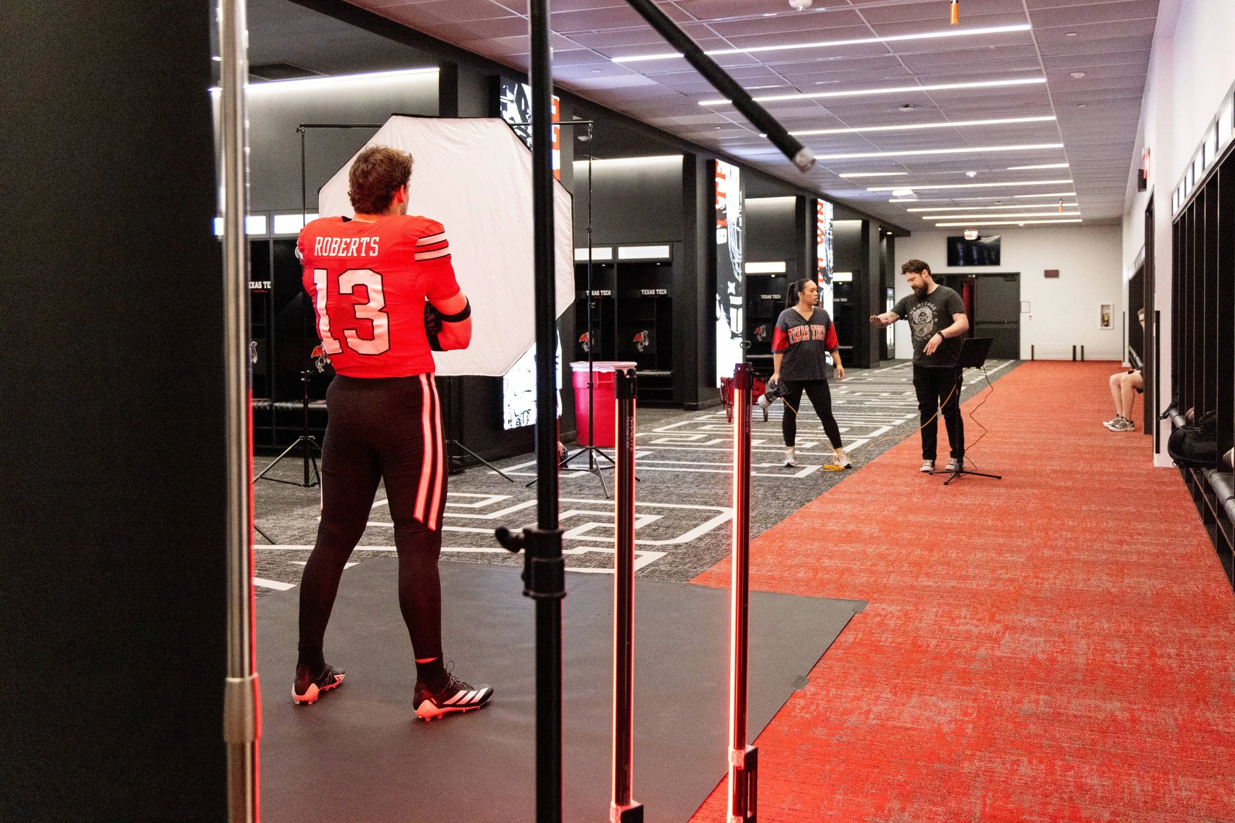 A person in a red sports jersey with the name 'Roberts' and the number '13' on the back, standing in a sports facility. There are two other individuals in the background, one with a camera and another in sportswear, near a backdrop with lighting equi