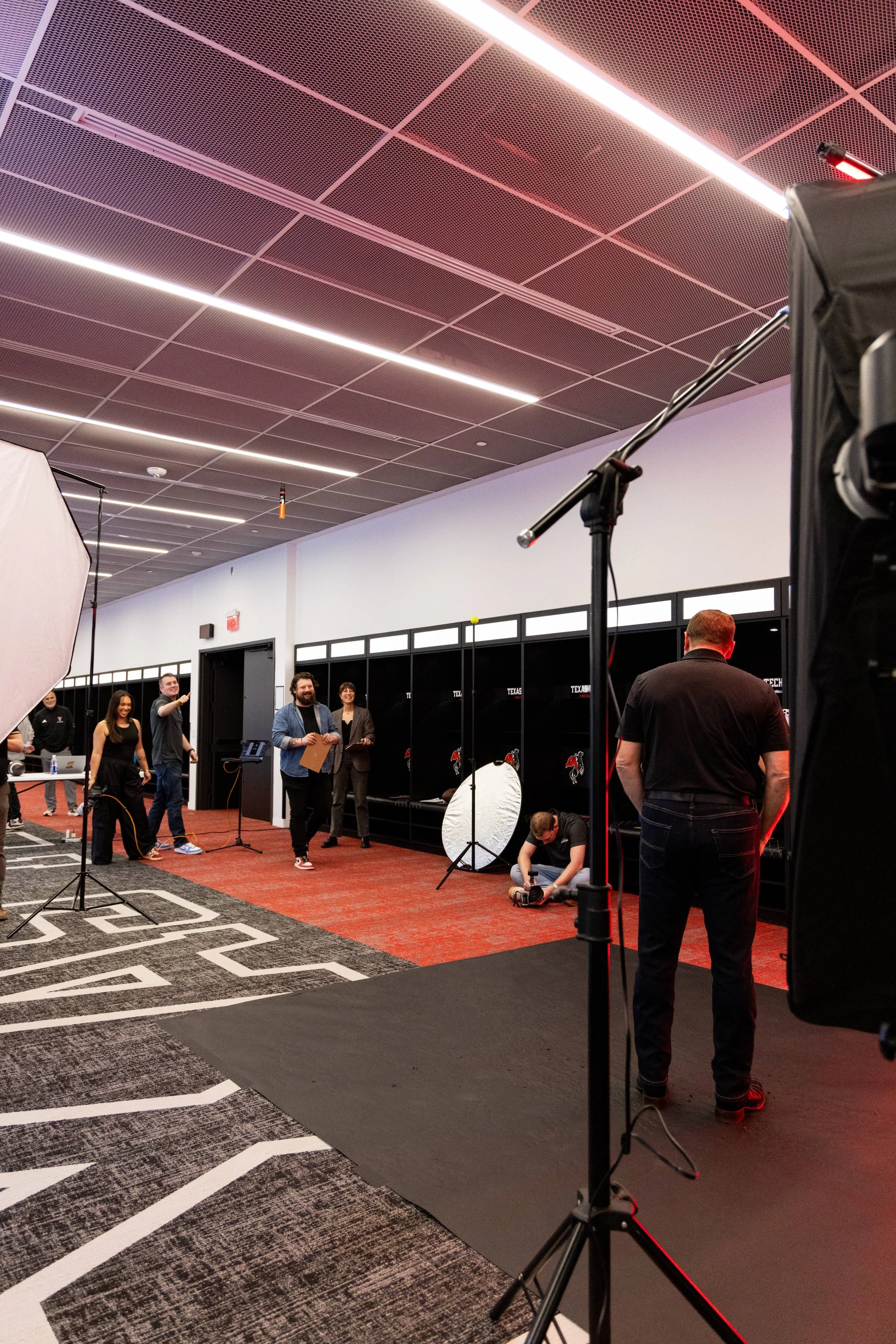 Group of people in a photo studio with photography equipment, cameras, and lights, preparing for a shoot in a room with black lockers, a red carpet, and a black backdrop.