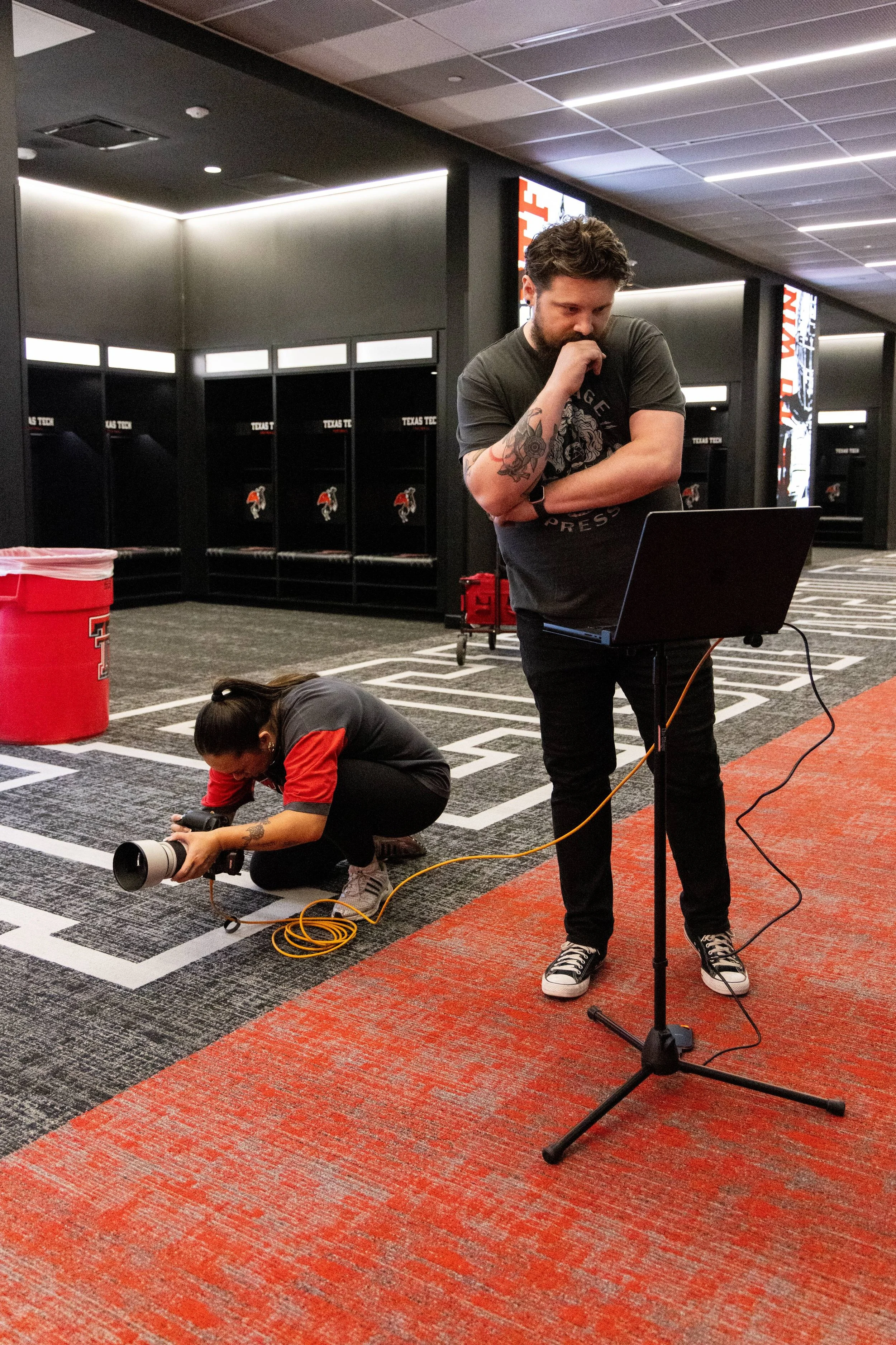 A person with tattoos on their arm is crouched on the floor, handling a camera with a large telephoto lens. Another person in a black t-shirt and sneakers is standing nearby, looking at a laptop on a stand. The background features lockers with sports