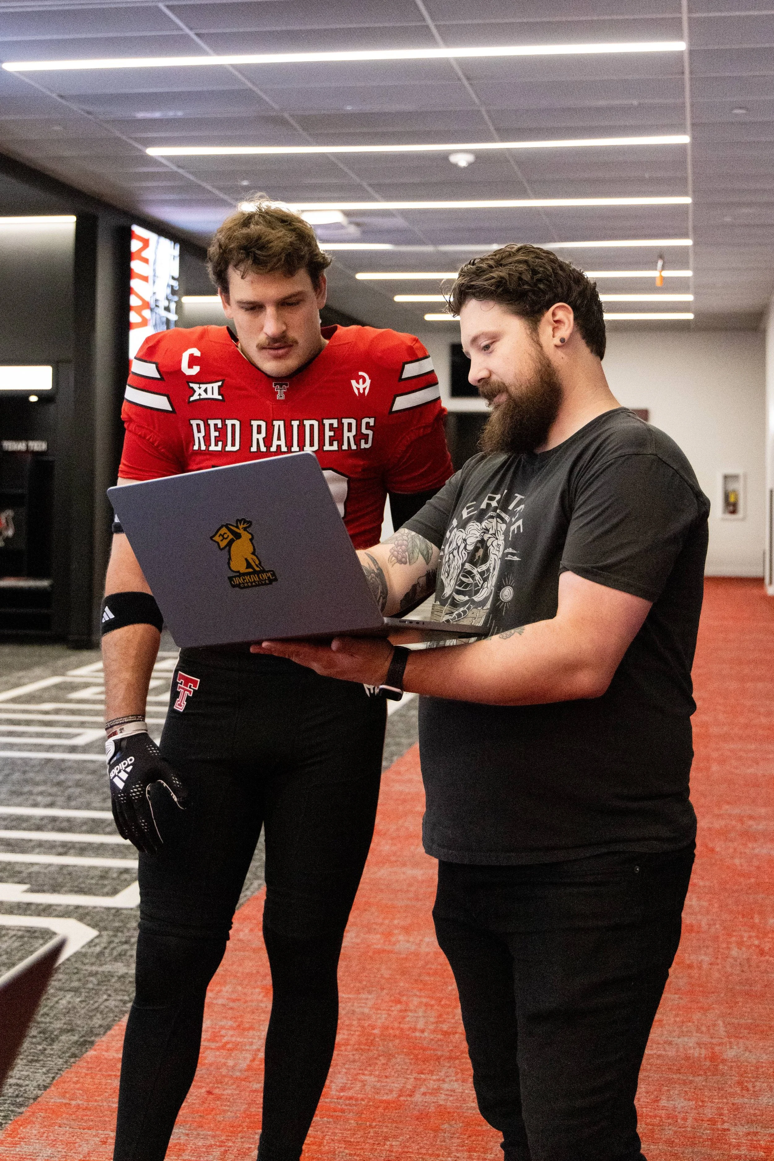 Two men standing in an indoor hallway looking at a laptop computer. One man is wearing a red football jersey and gloves, and the other man is dressed in a black T-shirt with tattoos on his arms.
