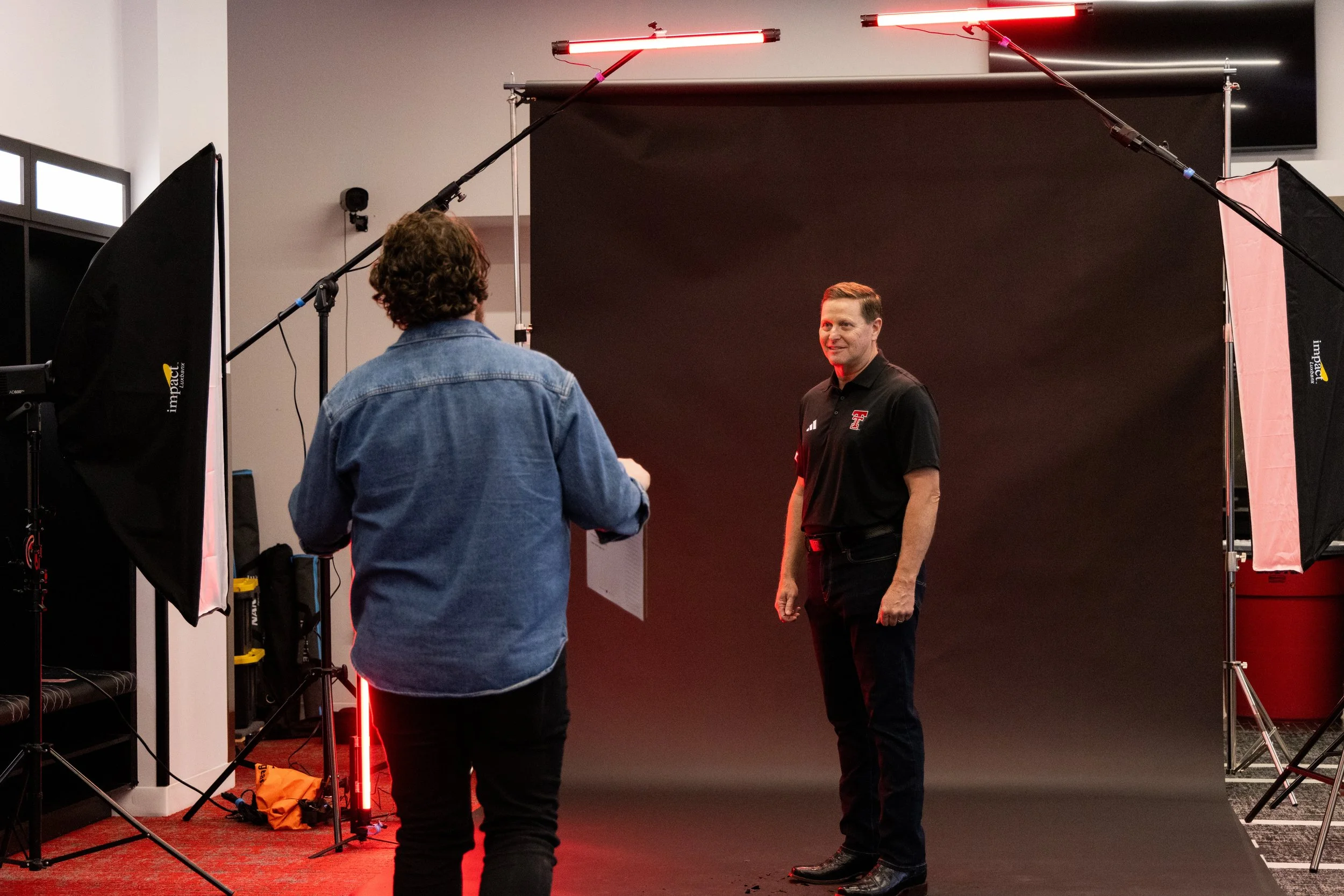 A photographer sets up a photoshoot with a man wearing a black polo shirt with a Texas Tech logo, standing in front of a dark backdrop, surrounded by studio lighting equipment.