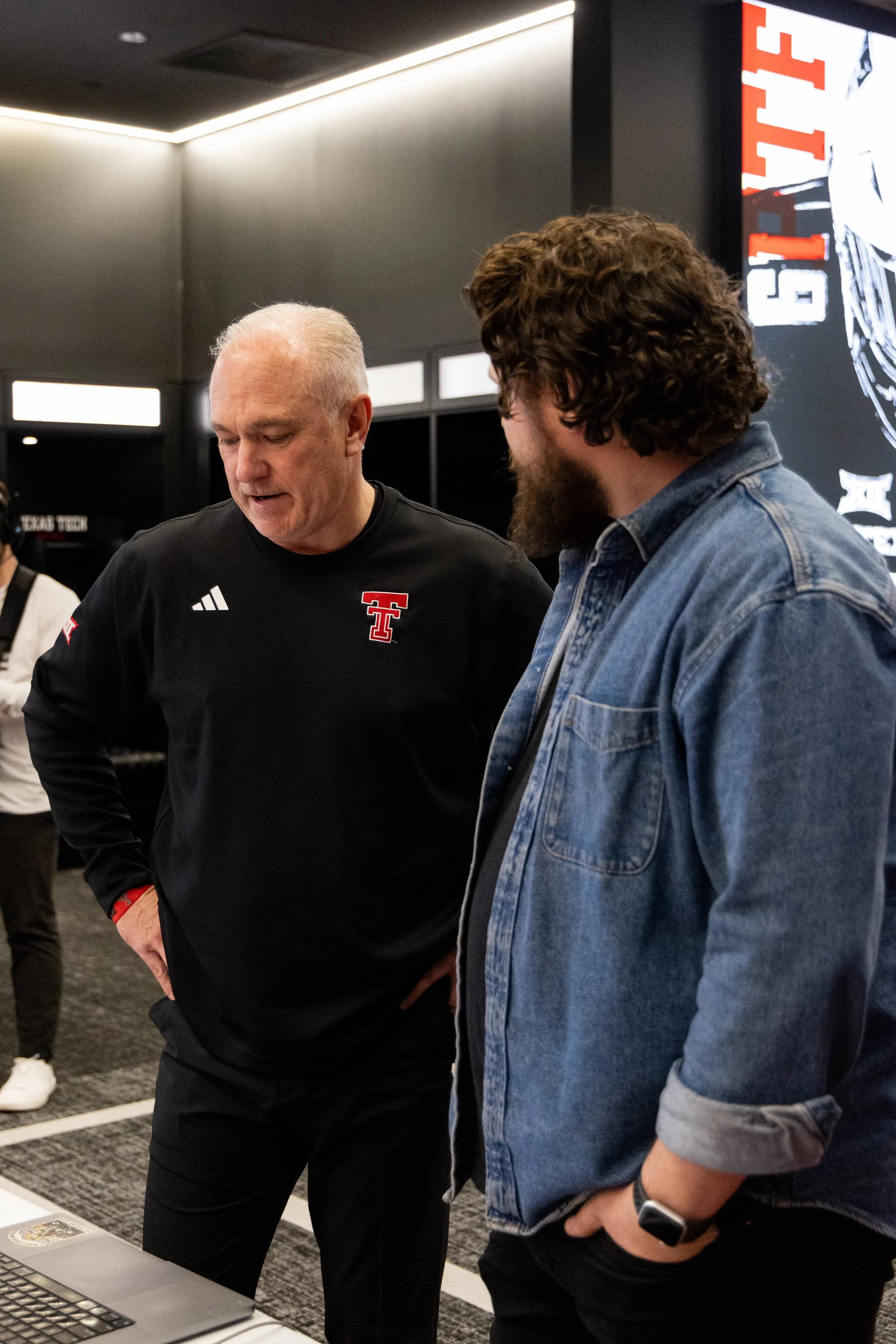 Two men are standing and looking at a laptop on a table. One man has gray hair and is wearing a black shirt with a logo on it, and the other man has curly brown hair, a beard, and is wearing a denim jacket.