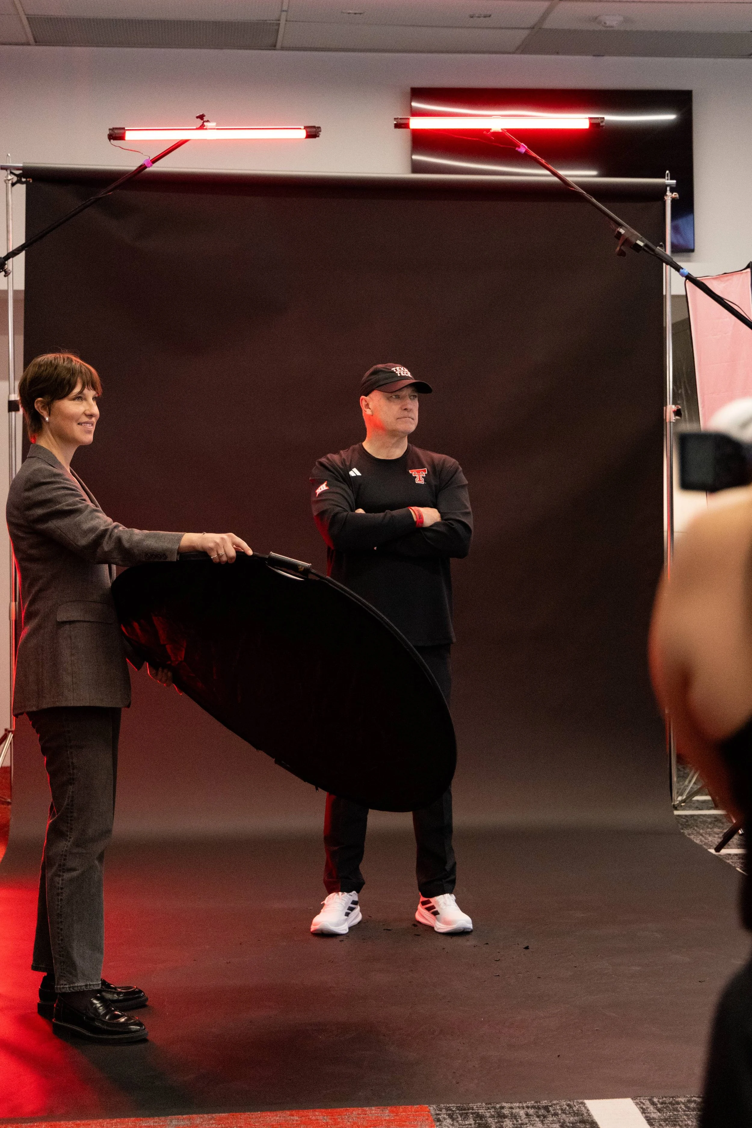 A woman holding a reflector and a man with crossed arms standing in front of a black backdrop with red lighting, likely during a photoshoot or interview.