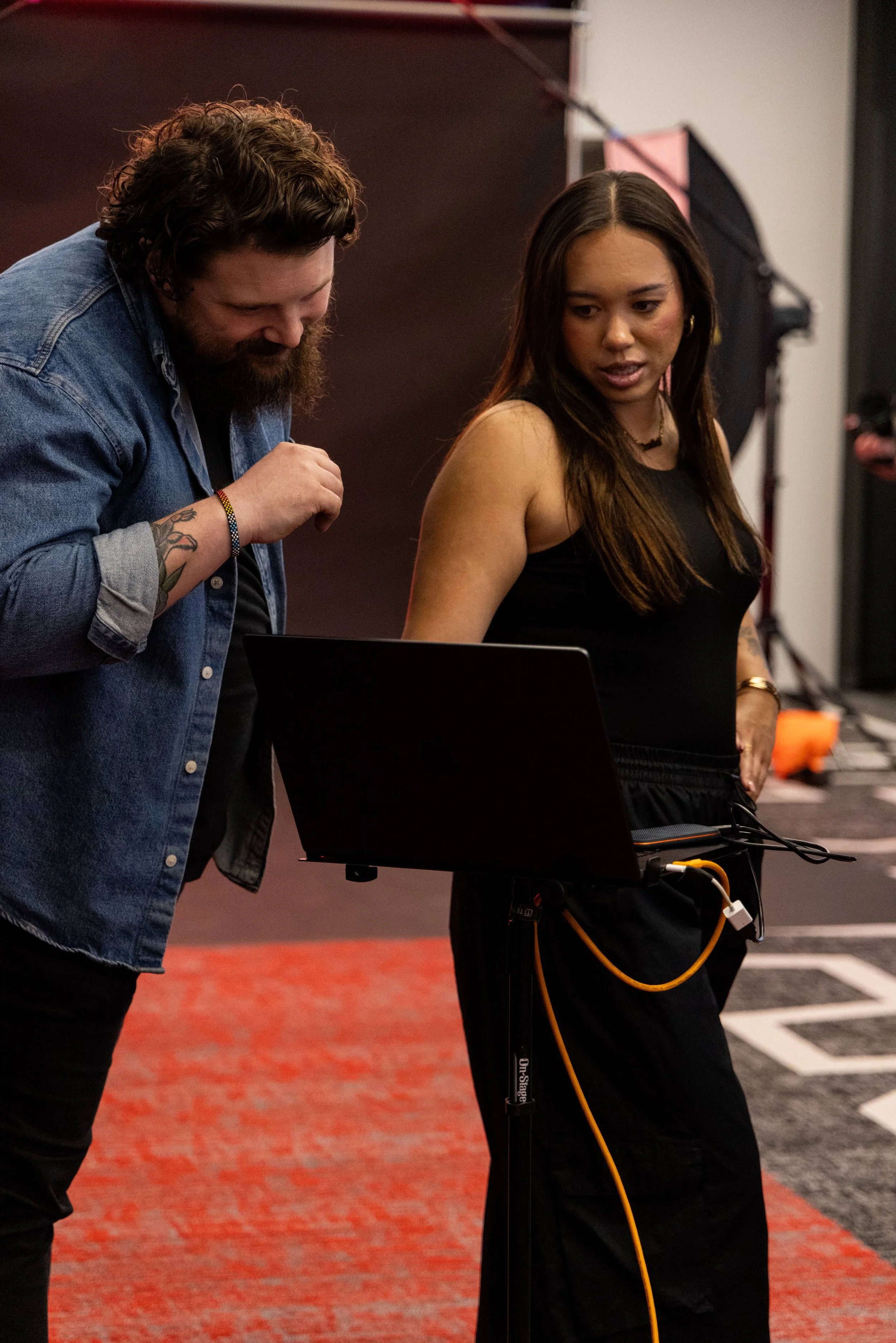 Two people are standing beside a laptop computer with a black screen. The person on the left is a man with curly brown hair, to a beard, wearing a denim shirt, and looking at the laptop screen. The person on the right is a woman with long dark brown 