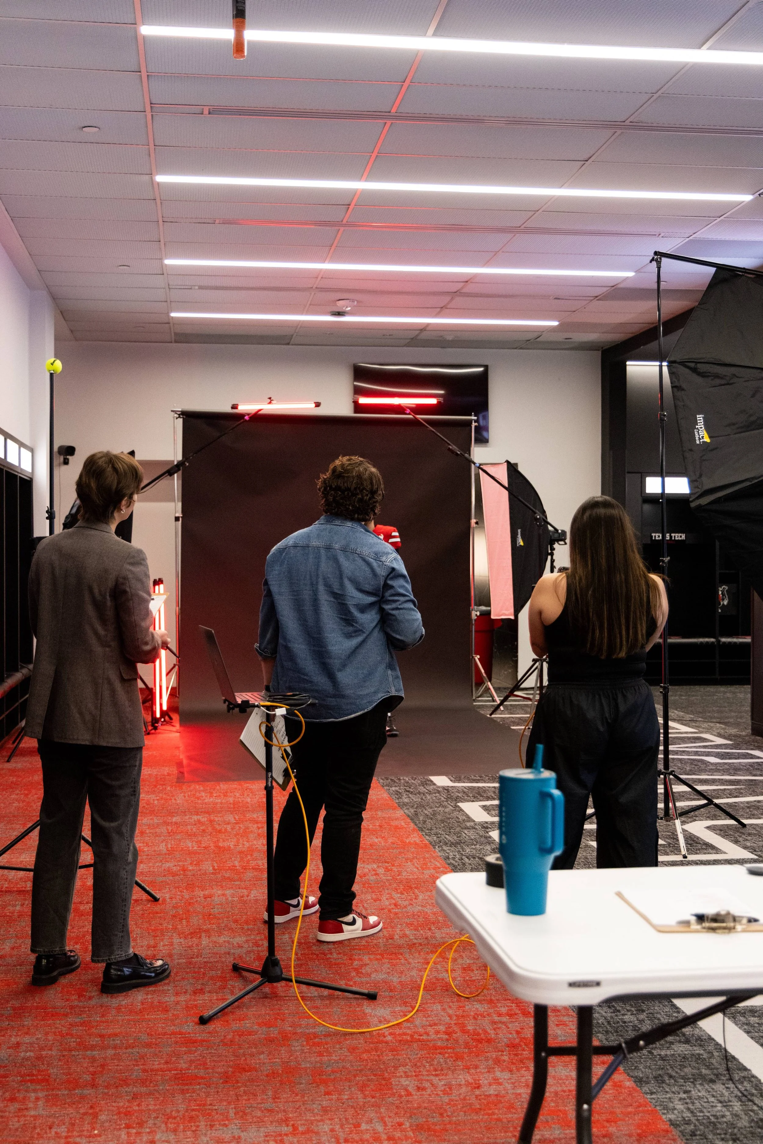 People working in a photography studio with lighting and backdrop equipment, some facing away from the camera, and a small table with a blue tumbler in the foreground.