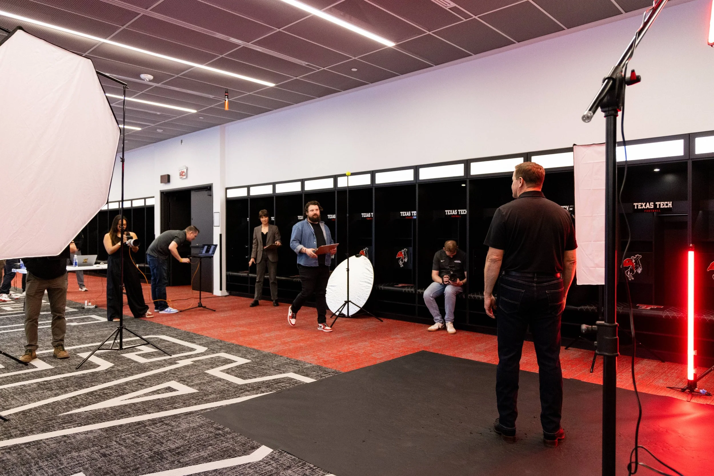Indoor sports facility with a backdrop of lockers labeled 'Texas Tech.' People are setting up a photo or video shoot with lighting equipment, photographers, and a backdrop, possibly for a media event or interview.