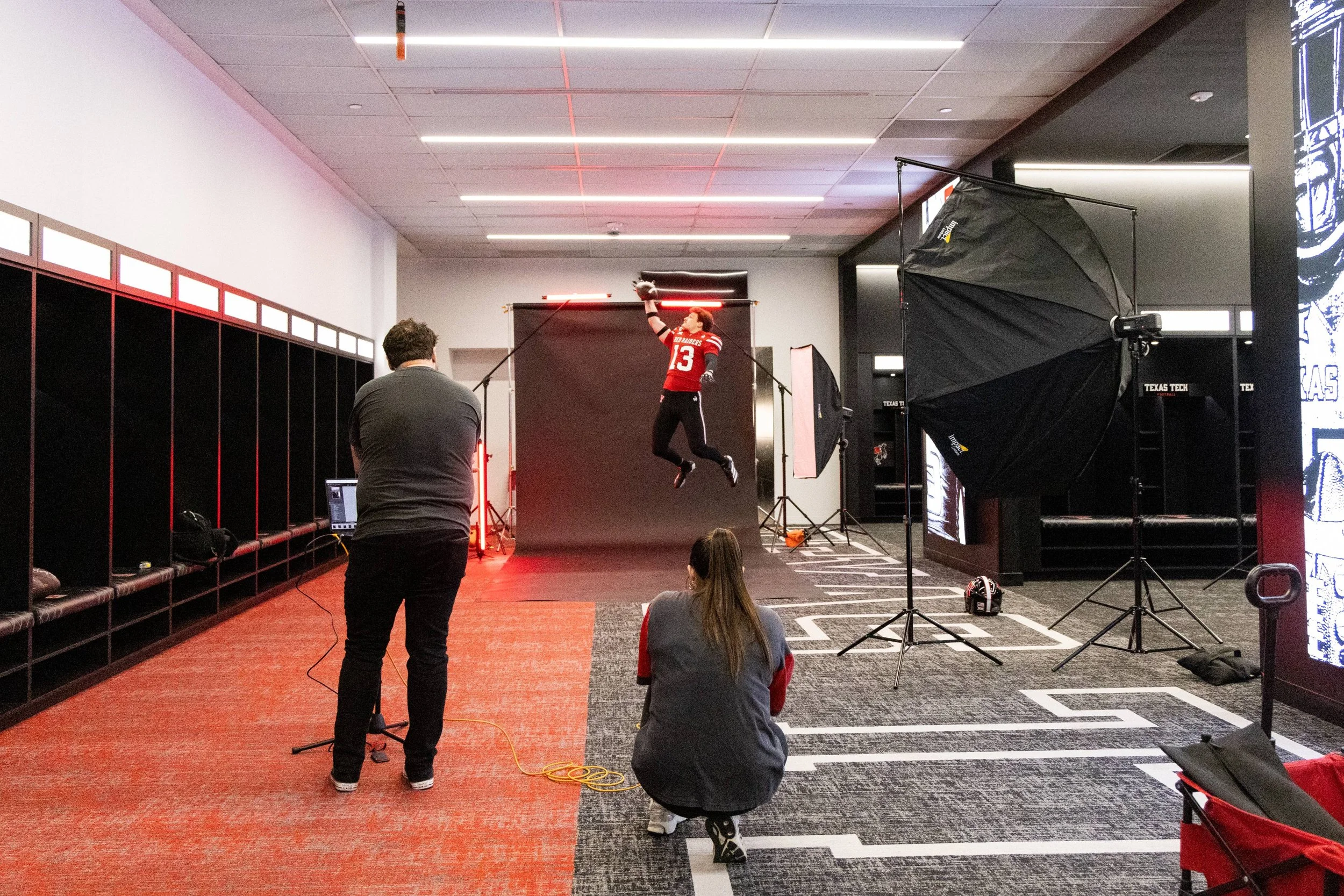 A person in athletic clothing, wearing a jersey with the number 13, is jumping in the air with a football, captured during a photoshoot in a studio with lighting equipment and a backdrop.