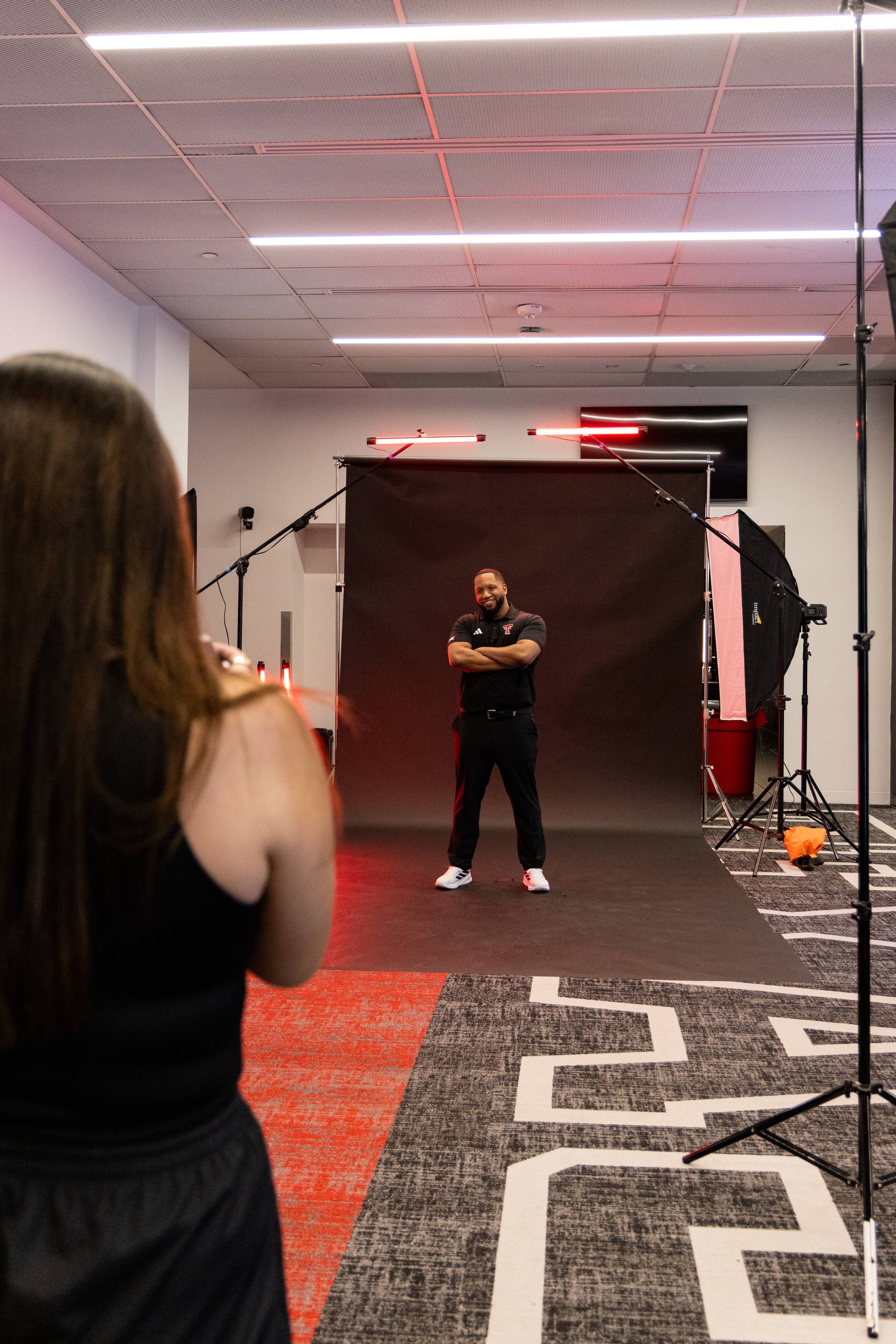 A man standing in front of a black backdrop during a photo shoot, smiling with his arms crossed. Studio lighting and equipment are visible, with a person in the foreground watching.