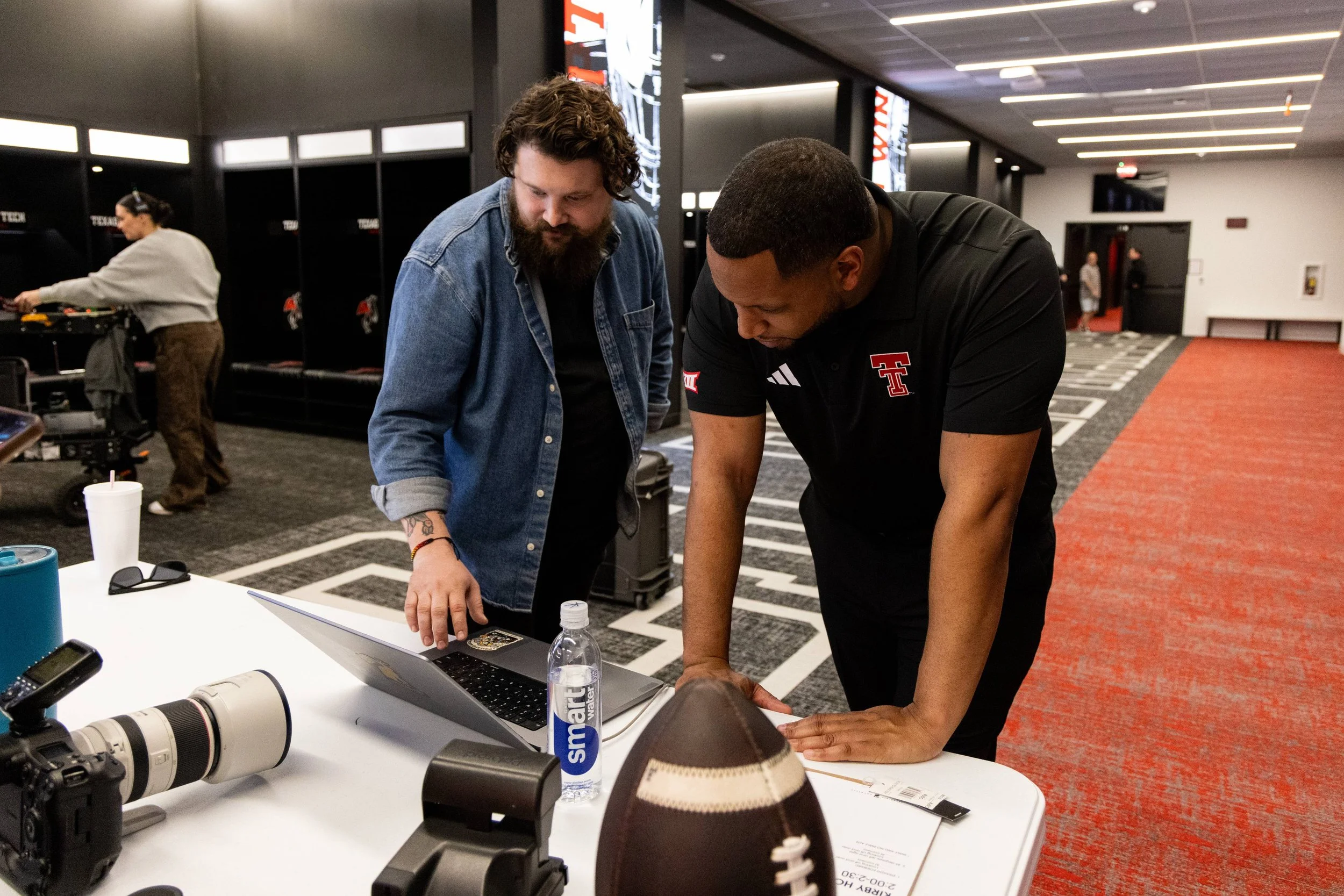 Two men are standing at a table with various items, including a large camera lens, a water bottle, a hat, a notebook, and electronic devices, in a room with black and white walls, digital screens, and a few other people in the background.