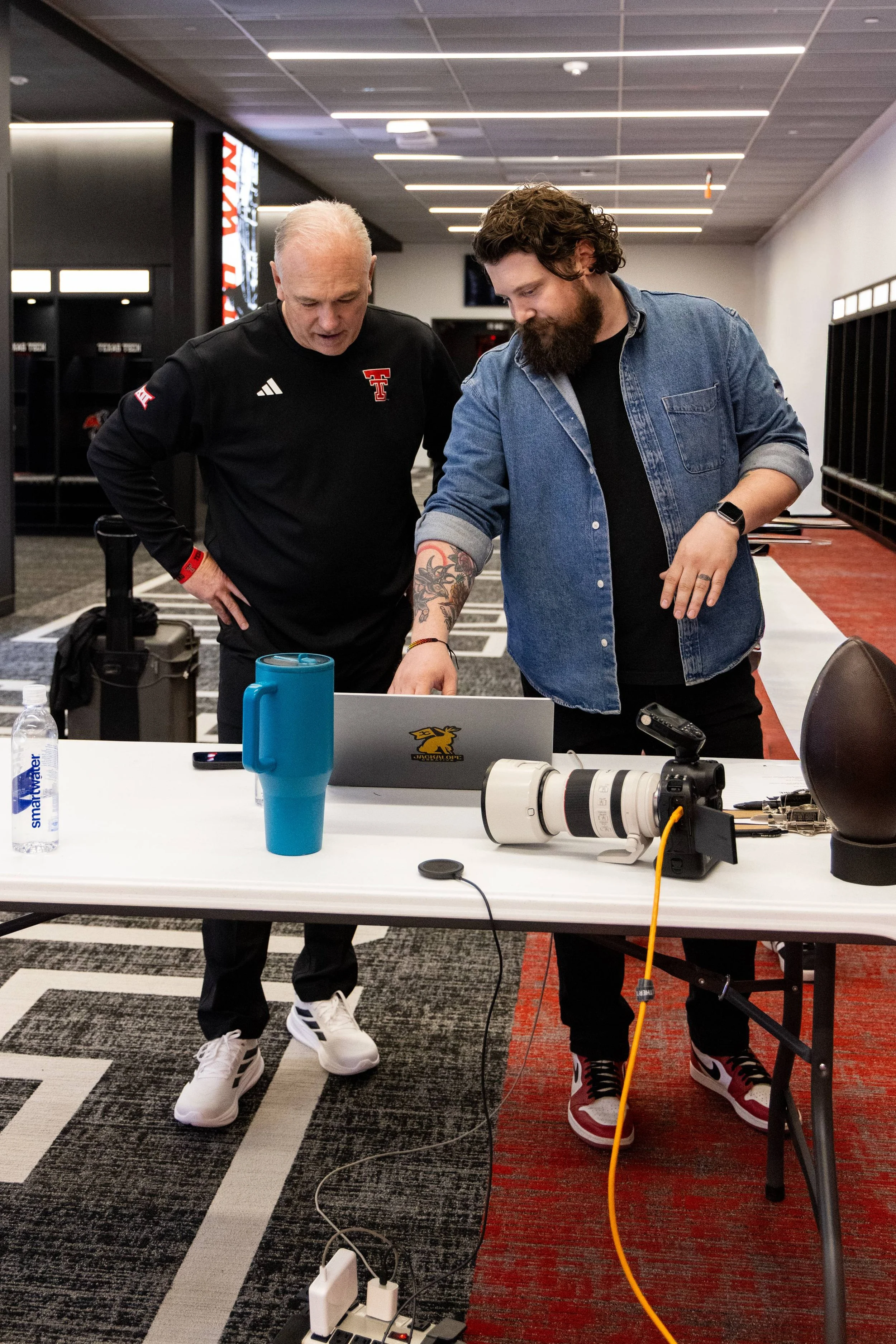 Two men are standing at a table, looking at a laptop with a dragon logo. The man on the left has short gray hair and is wearing a black long-sleeved shirt with sports logos. The man on the right has curly brown hair, a beard, and is wearing a denim s