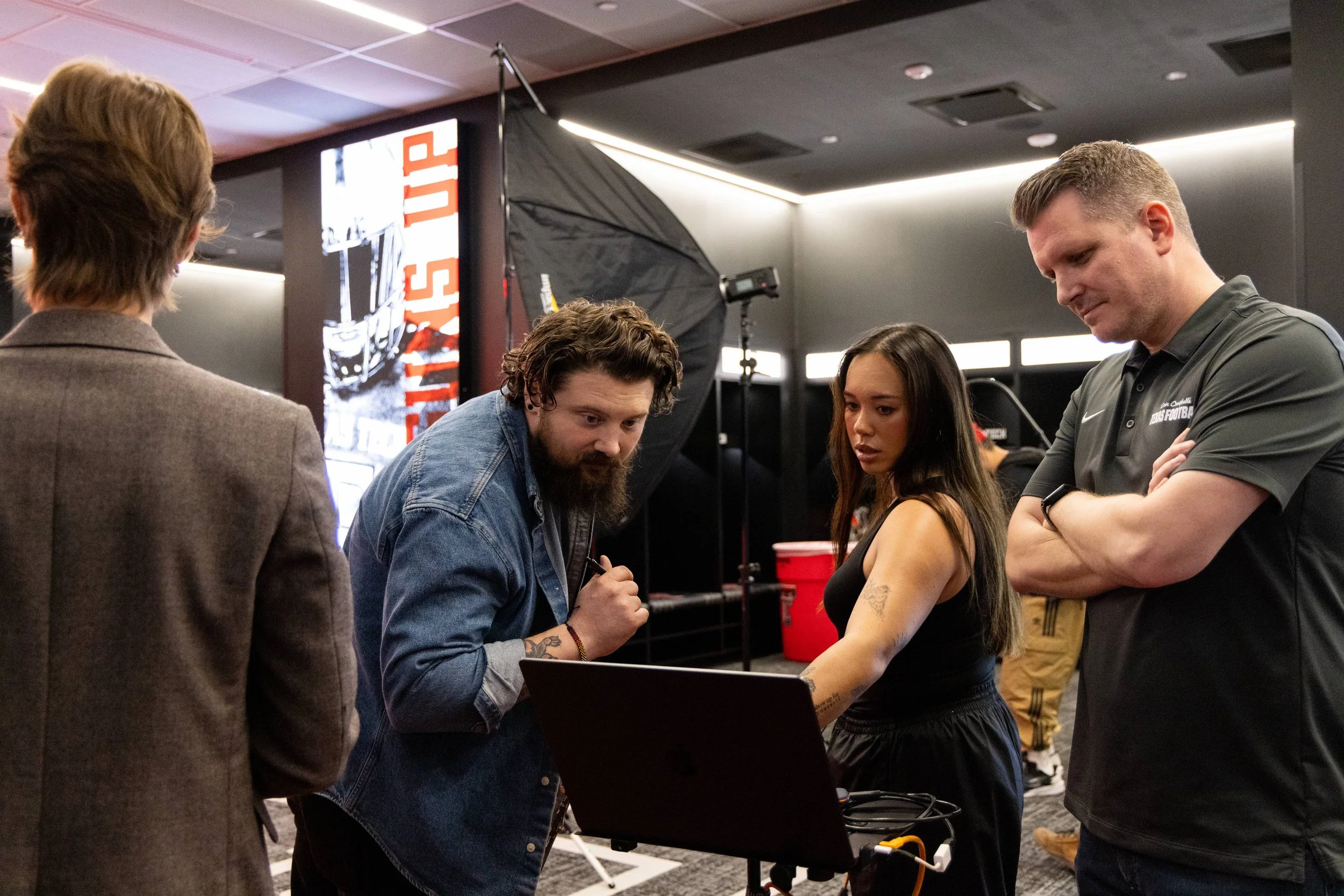 Five people are gathered around a table in a room with equipment and a large digital screen in the background. One person is signing an arm tattoo of a woman, while the others watch and stand nearby.