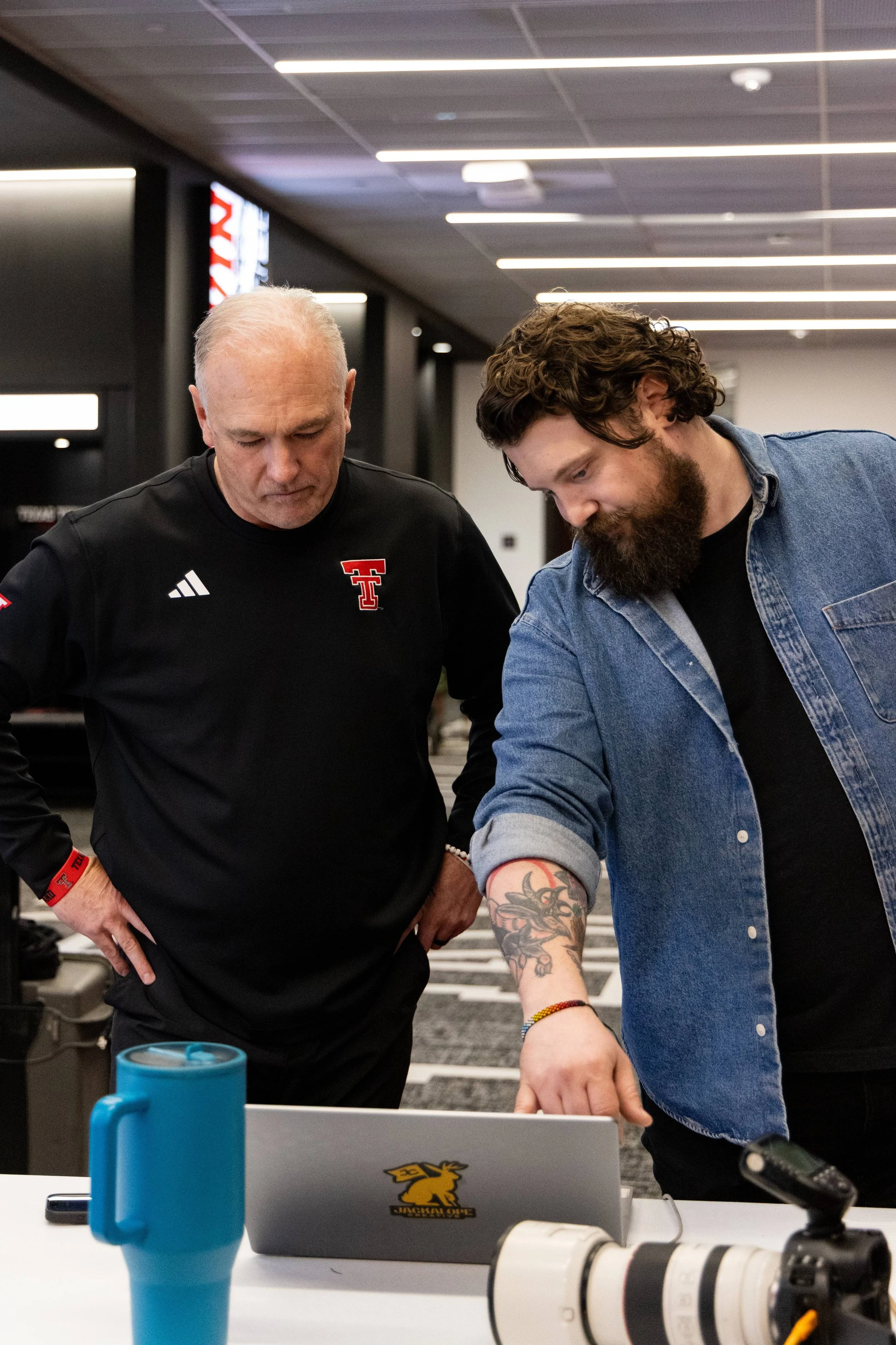 Two men working together at a table with various electronic equipment, including a camera and a blue tumbler, in a modern indoor space with fluorescent lighting.