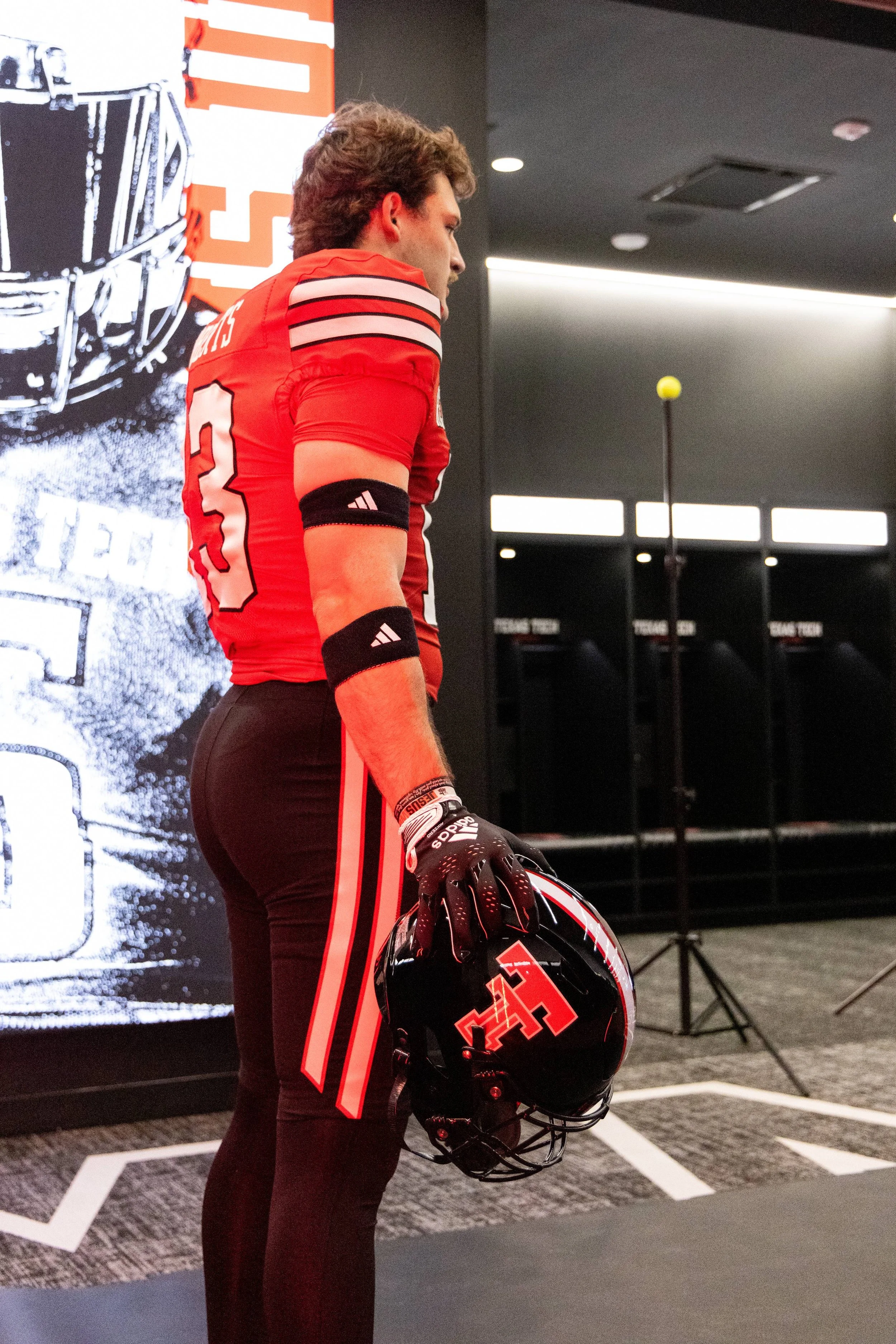 A male football player wearing a red and black uniform holding a black helmet with a red logo and a yellow football in the background.