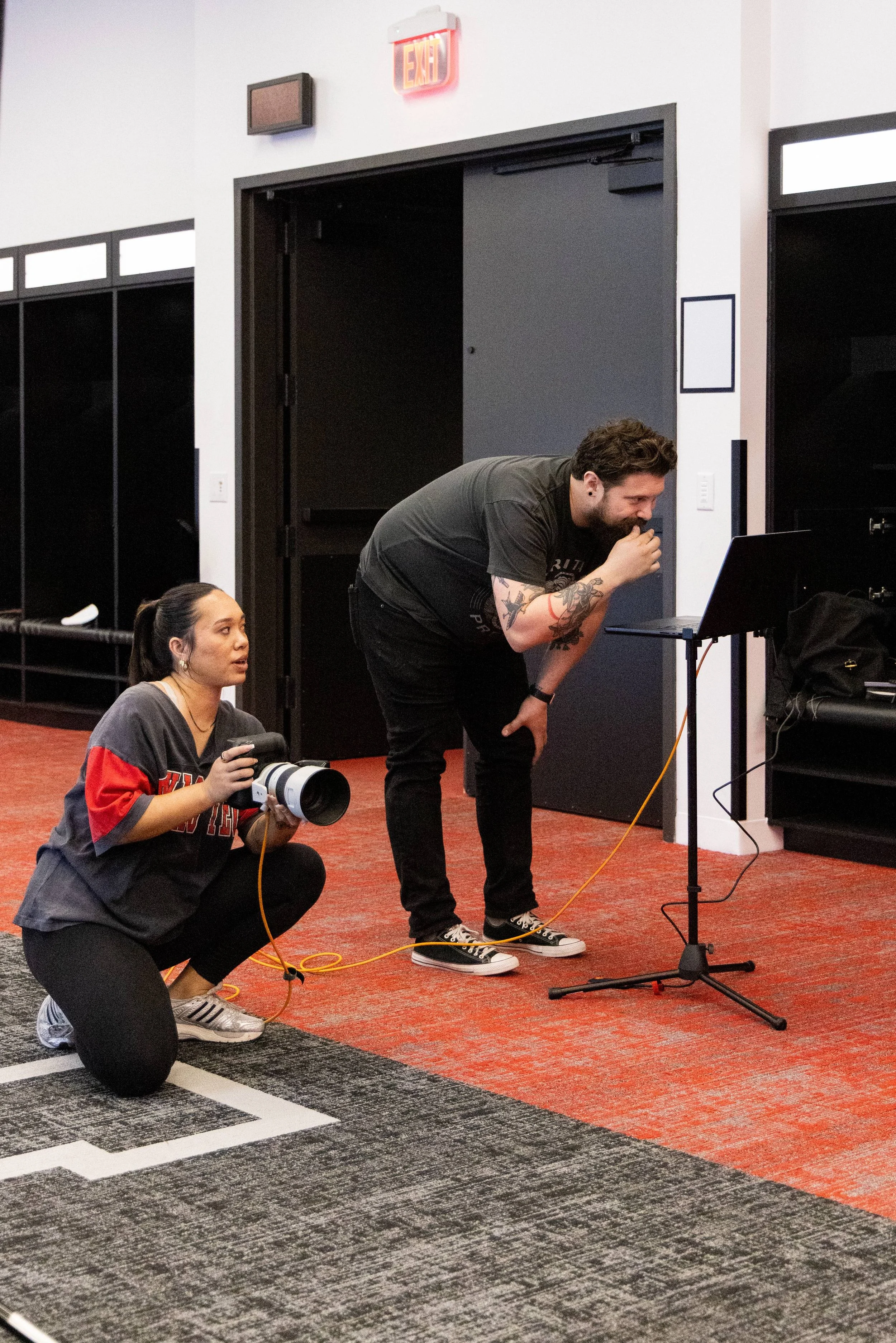 A woman squatting on a black platform with a camera around her neck, holding a wire. A man with tattoos and dark hair, leaning forward and looking at a laptop on a stand, appears to be explaining something. They are in an indoor space with black lock