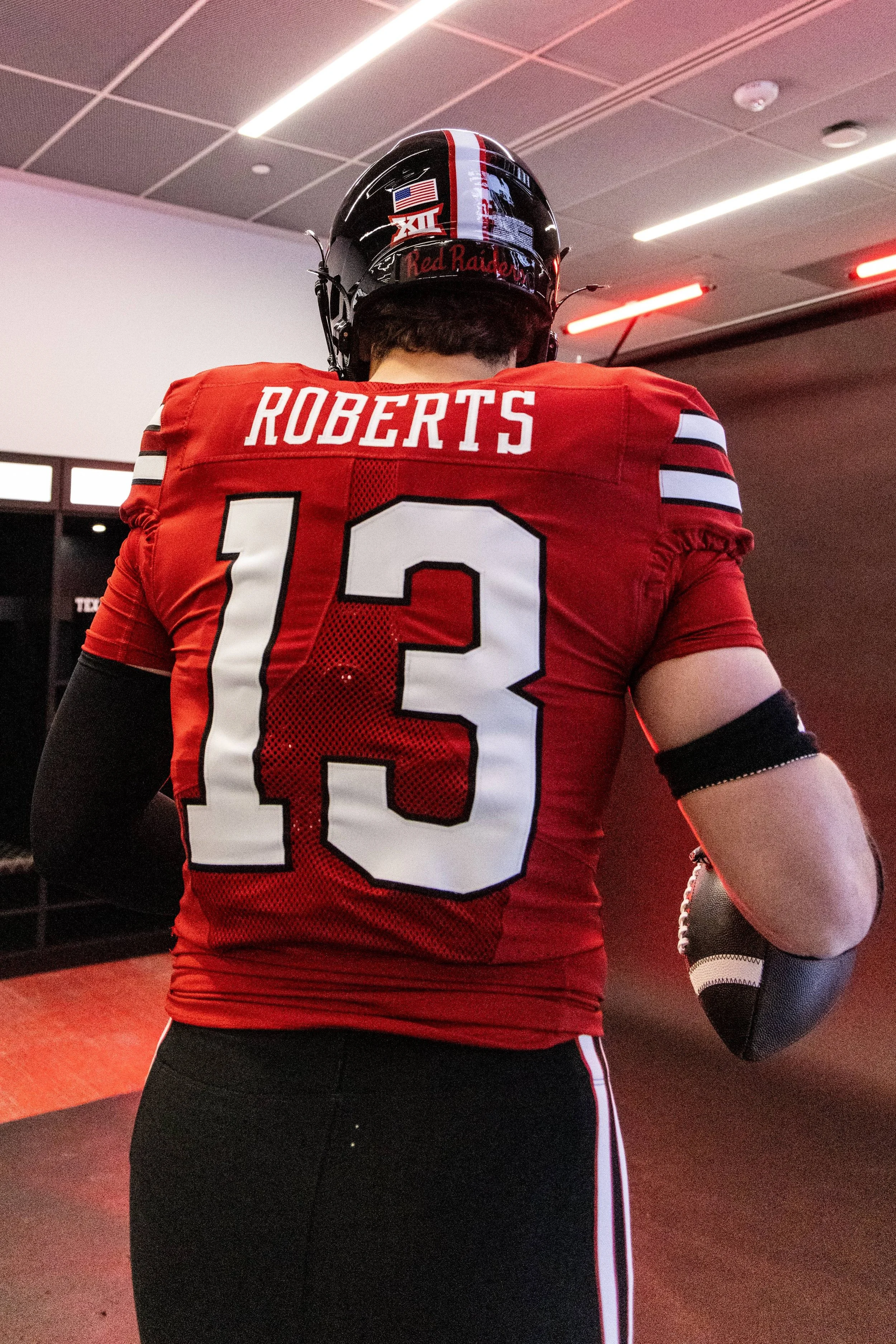 Back view of a football player in a red jersey with the number 13 and the name Roberts, standing indoors with a black helmet and a person holding a football in the background.