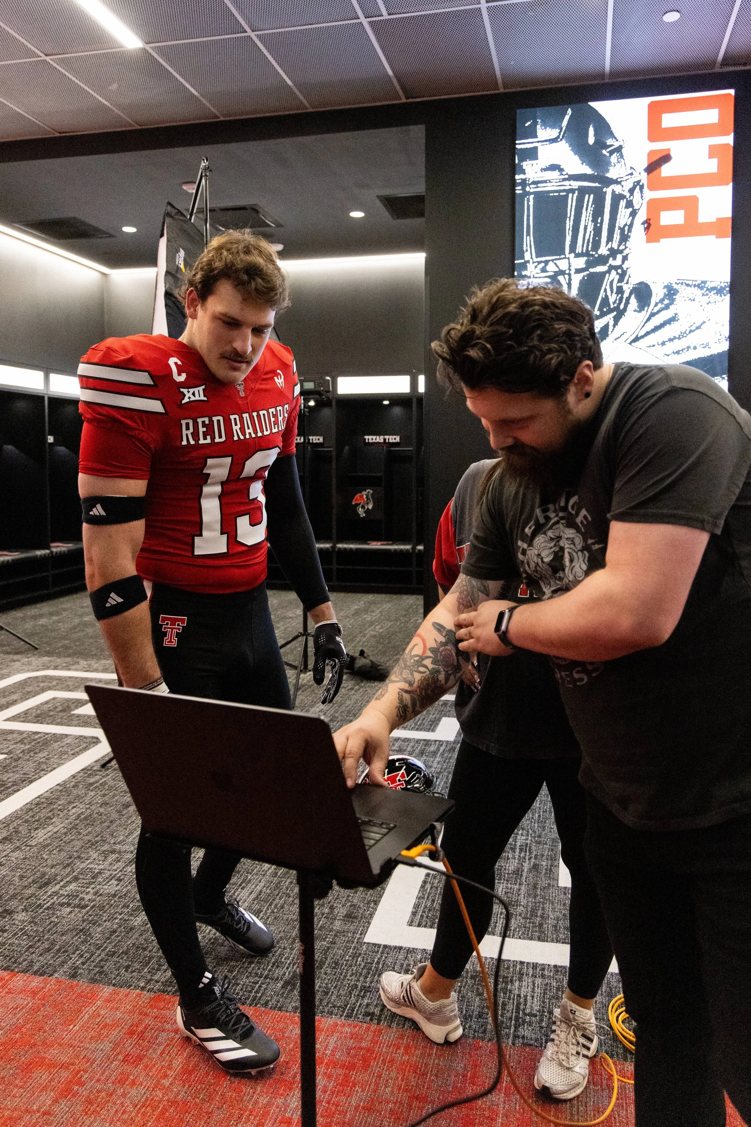 A football player in a red Texas Tech Red Raiders jersey and black pants is looking down at a laptop on a table, while another person with tattoos on their arm is also looking at the laptop. The background features a digital screen with the word 'PLA