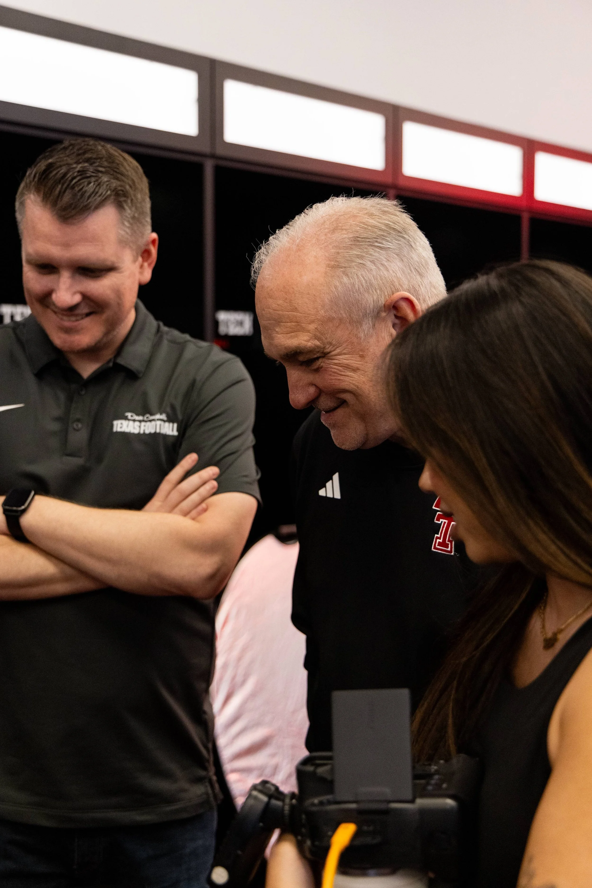 A group of people gathered around a computer, appearing to look at something on the screen. One person is wearing a dark shirt with a Texas Football logo, and another person is wearing a black shirt with an Adidas logo. The background shows lockers o