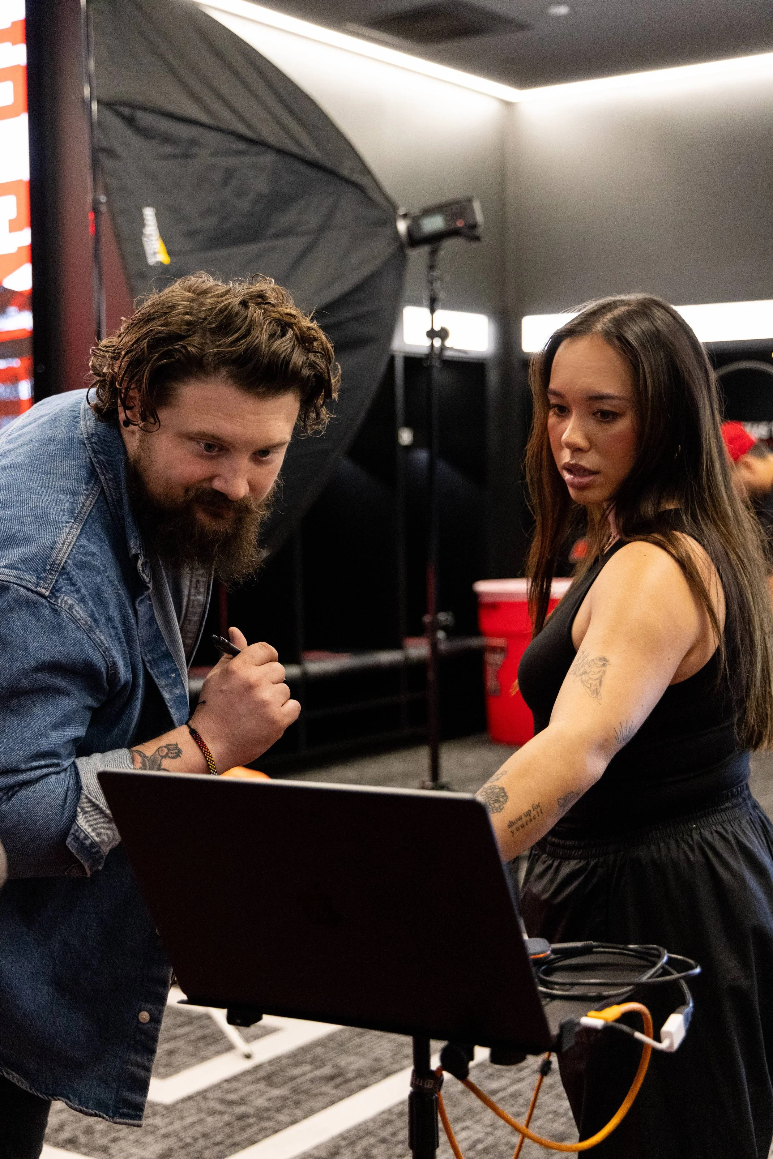 Two people working together in a photography studio, surrounded by equipment including a softbox, light stands, and a laptop. The woman has long dark hair, tattoos on her arms, and is looking at the man with a focused expression. The man has curly ha