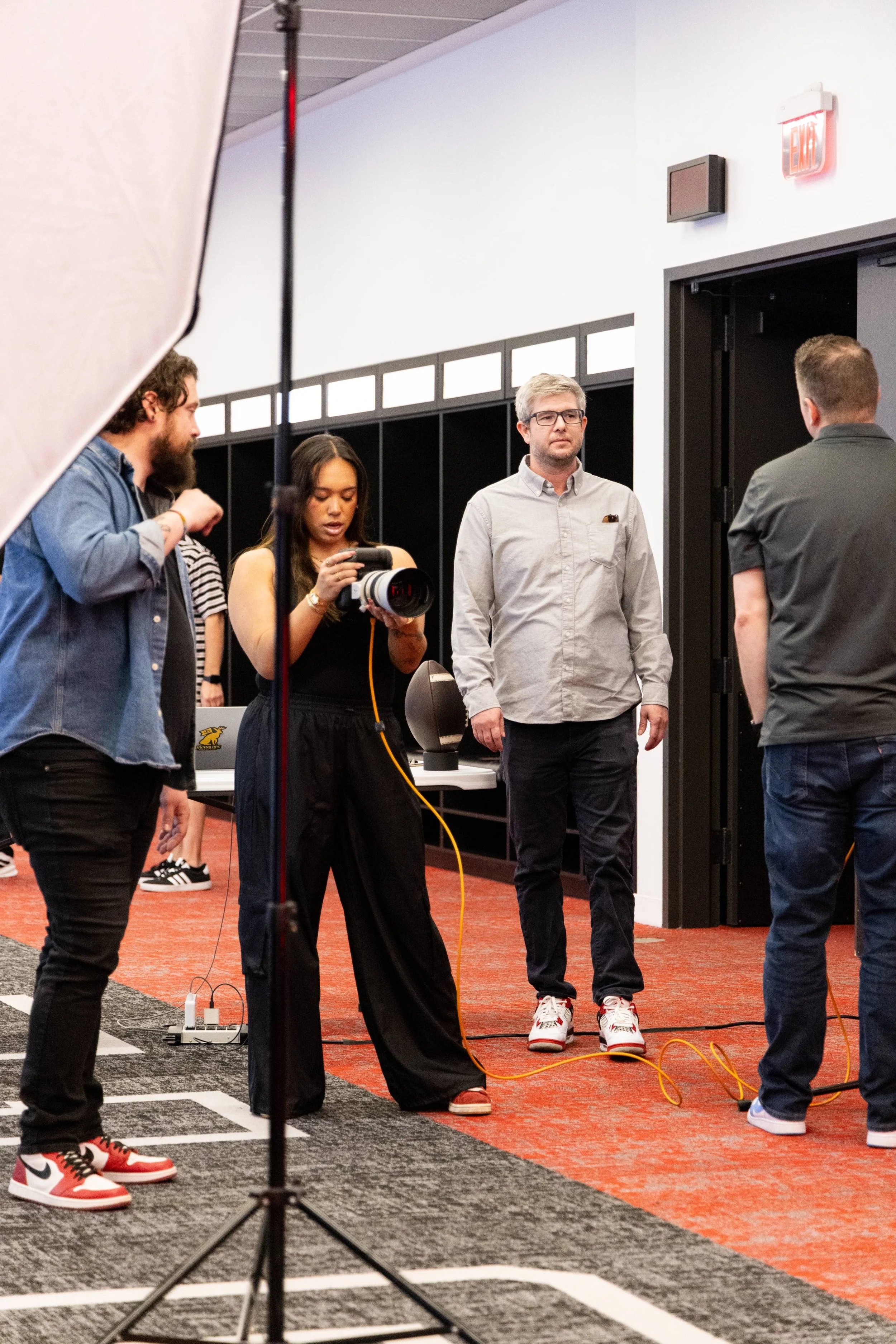 People in a room with photography equipment, standing on a grid-marked floor, possibly preparing for a photo or video shoot, with a black backdrop and a fluorescent exit sign visible.
