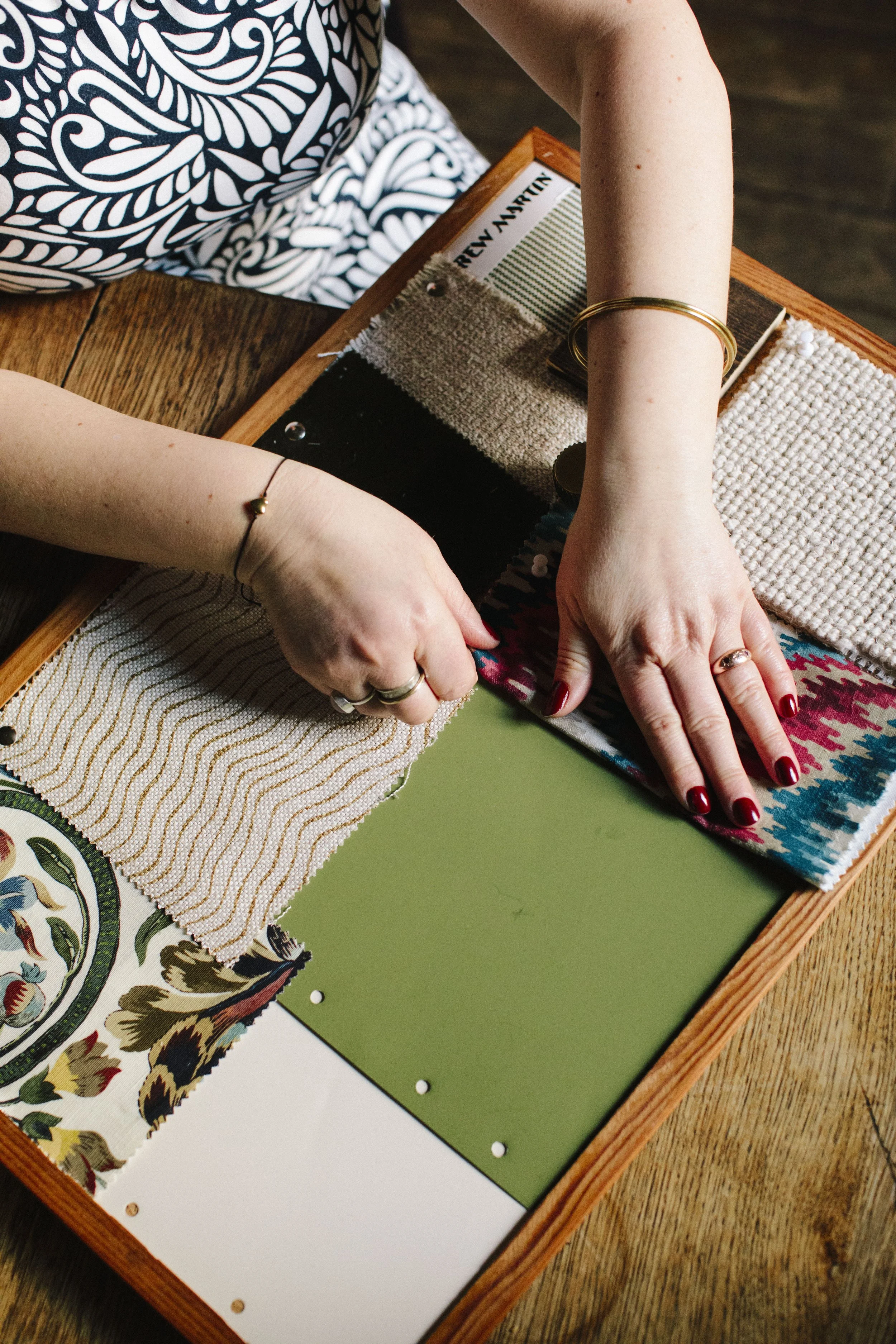 A pinboard with fabric and paint samples on it being smoothed by a pair of hands with red painted nails belonging to a white woman who is an interior designer