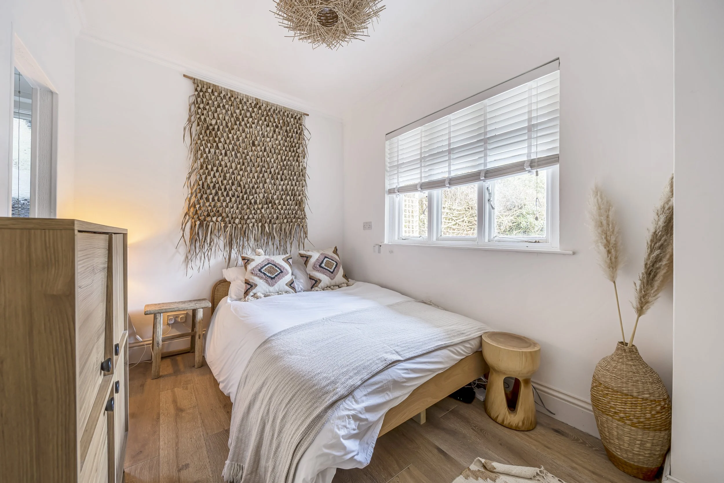 A bedroom with white painted walls, a double bed dressed with a white duvet and brown and black pillows with a diamond design. The room has lots of texture with a wicker lightshade, rattan vase on the floor rough wood stool used as a bedside table