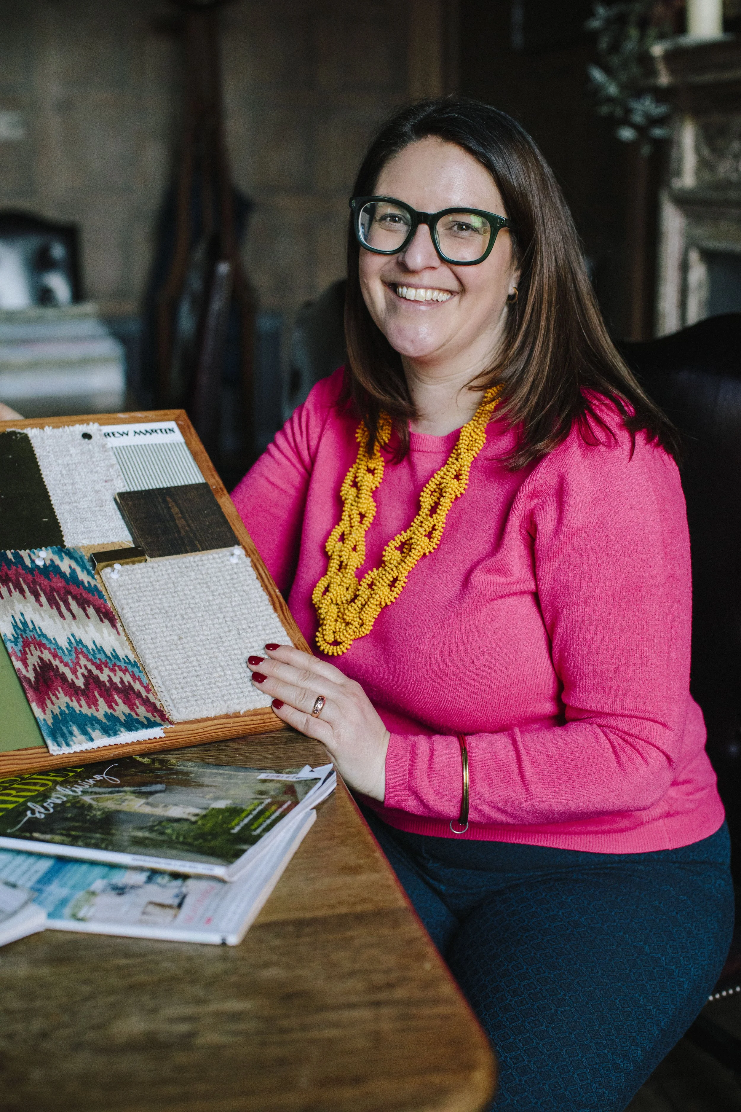 A white woman with brown hair and green glasses smiling at the camera. She is sat at a wooden table holding a interior design moodboard. She is dressed in a pink top with a yellow necklace and blue trousers