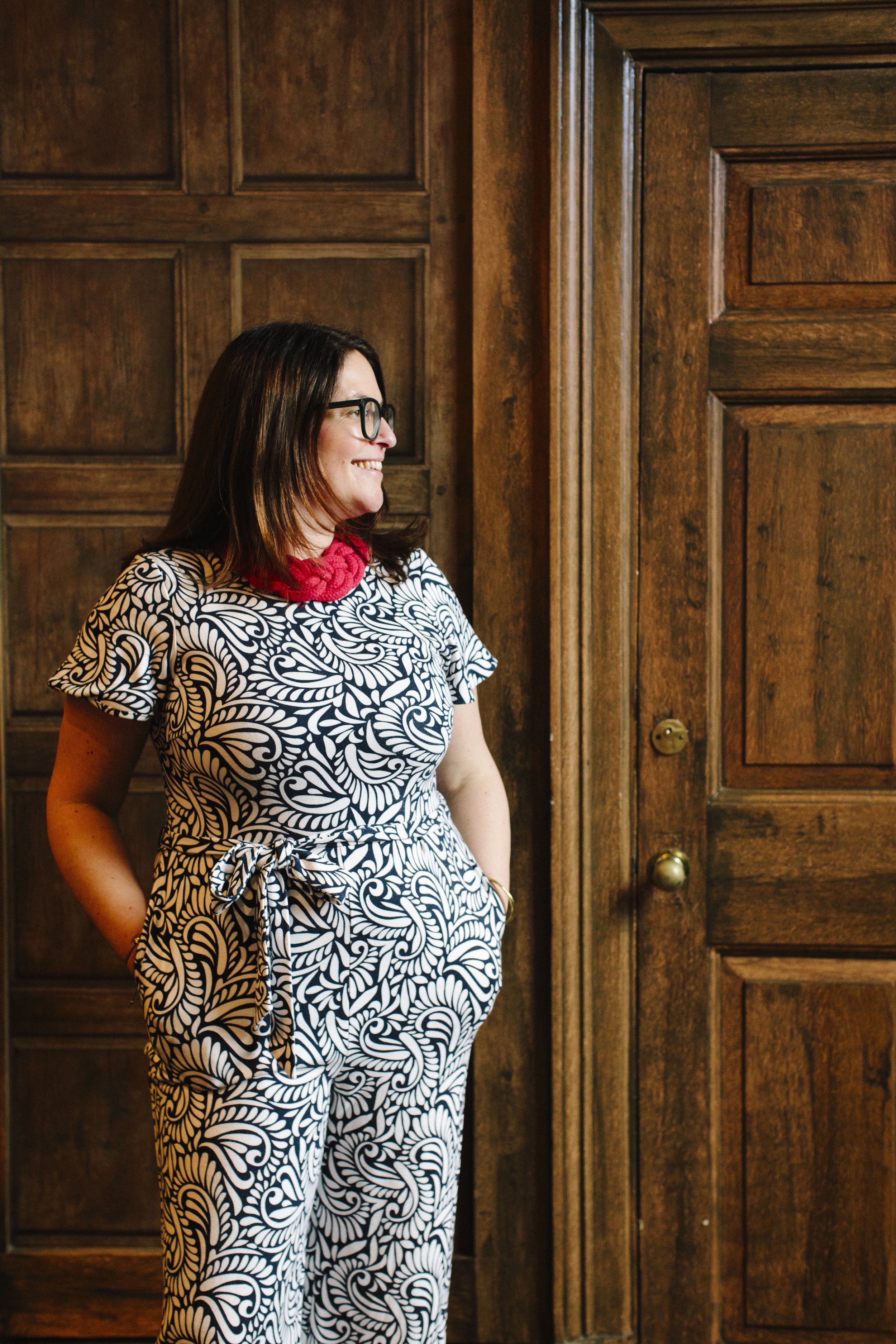 A white woman with brown hair wearing a patterned blue and white jumpsuit with a red necklace is stood in front of a pair of wooden double doors