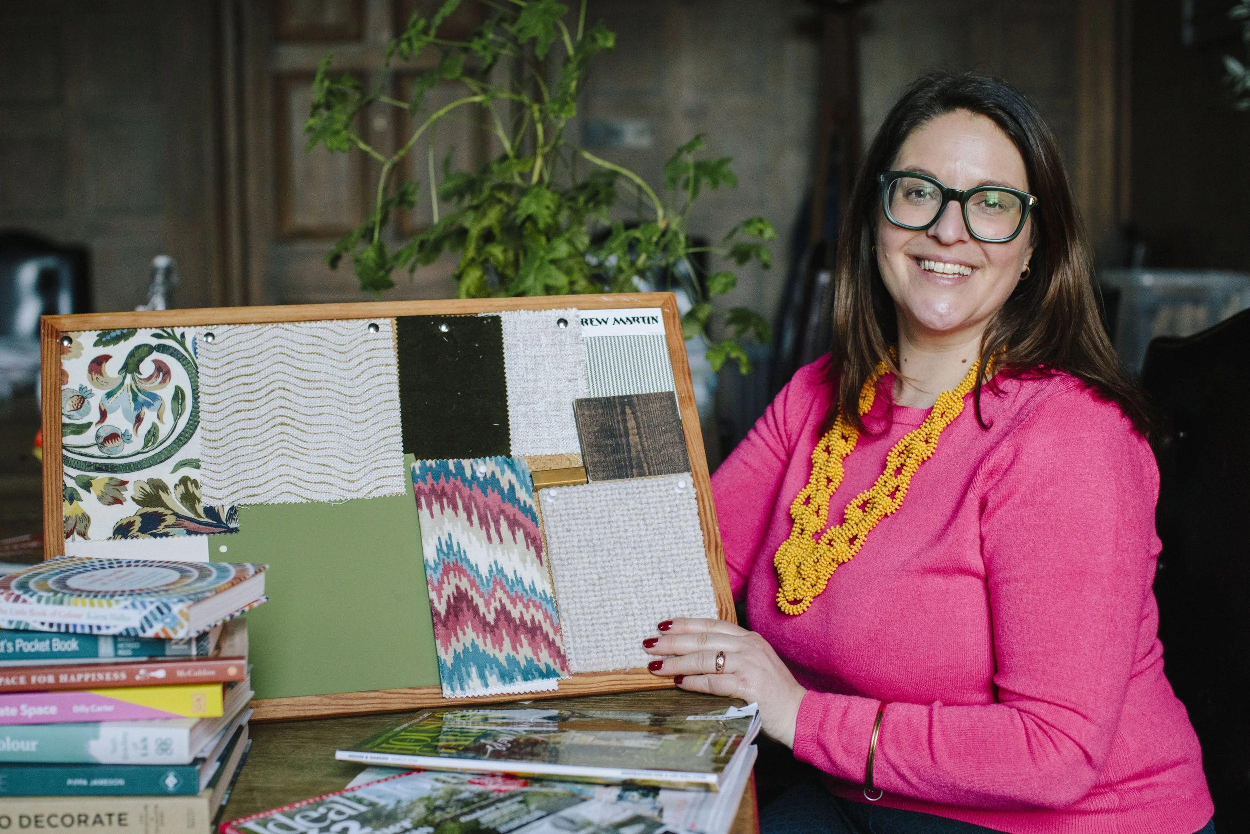A white female interior designer sits at a table looking directly at the camera and smiling. She is wearing a pink jumper with a yellow necklace and she wears green rimmed glasses. She is holding a moodboard with fabric and paint samples pinned to it