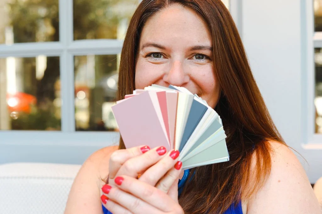 Becky Harrison, interior designer holding a fan deck of Fenwick & Tilbrook paint samples. She is holding the colourful fan deck over her mouth but you can tell from her eyes that she is smiling