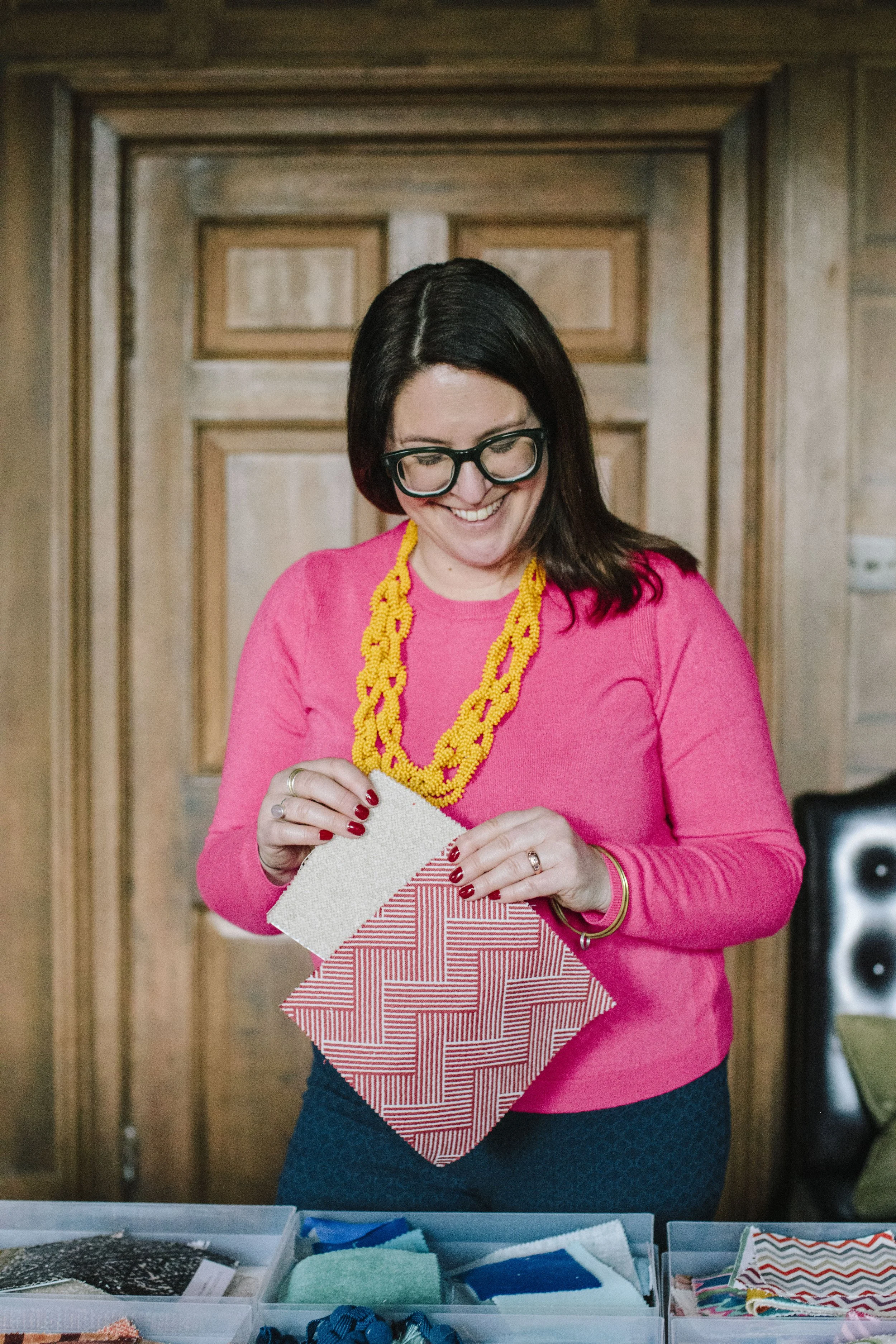 A white woman with brown hair smiles down at trays of fabric samples. She is dressed in a hot pink jumper with a large yellow necklace. She is smiling and in her hands has two fabric samples that she is looking down at