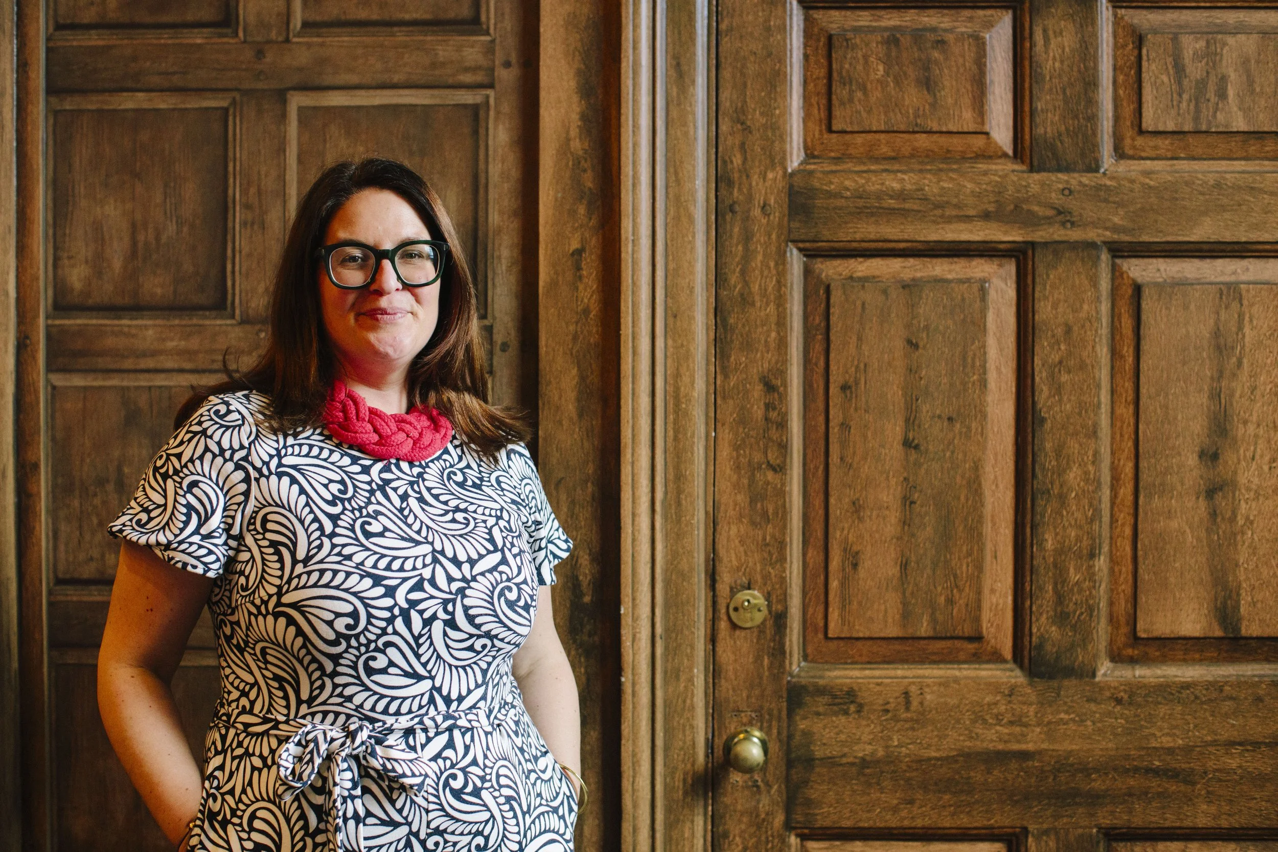 An interior designer who is female with white skin, brown hair and green glasses stands in front of a wooden panelled room and is looking confidently directly at the camera