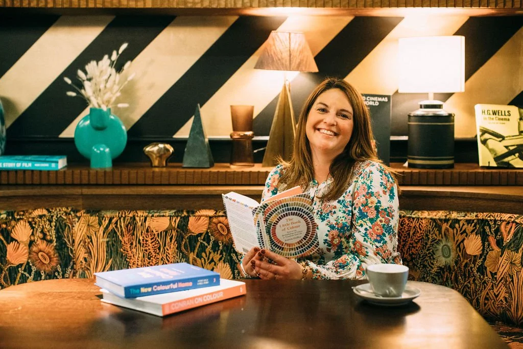 A white woman with brown hair smiling whilst reading a book about colour. She is sat on a velvet patterned sofa with a black and white geometirc wall paper in the background