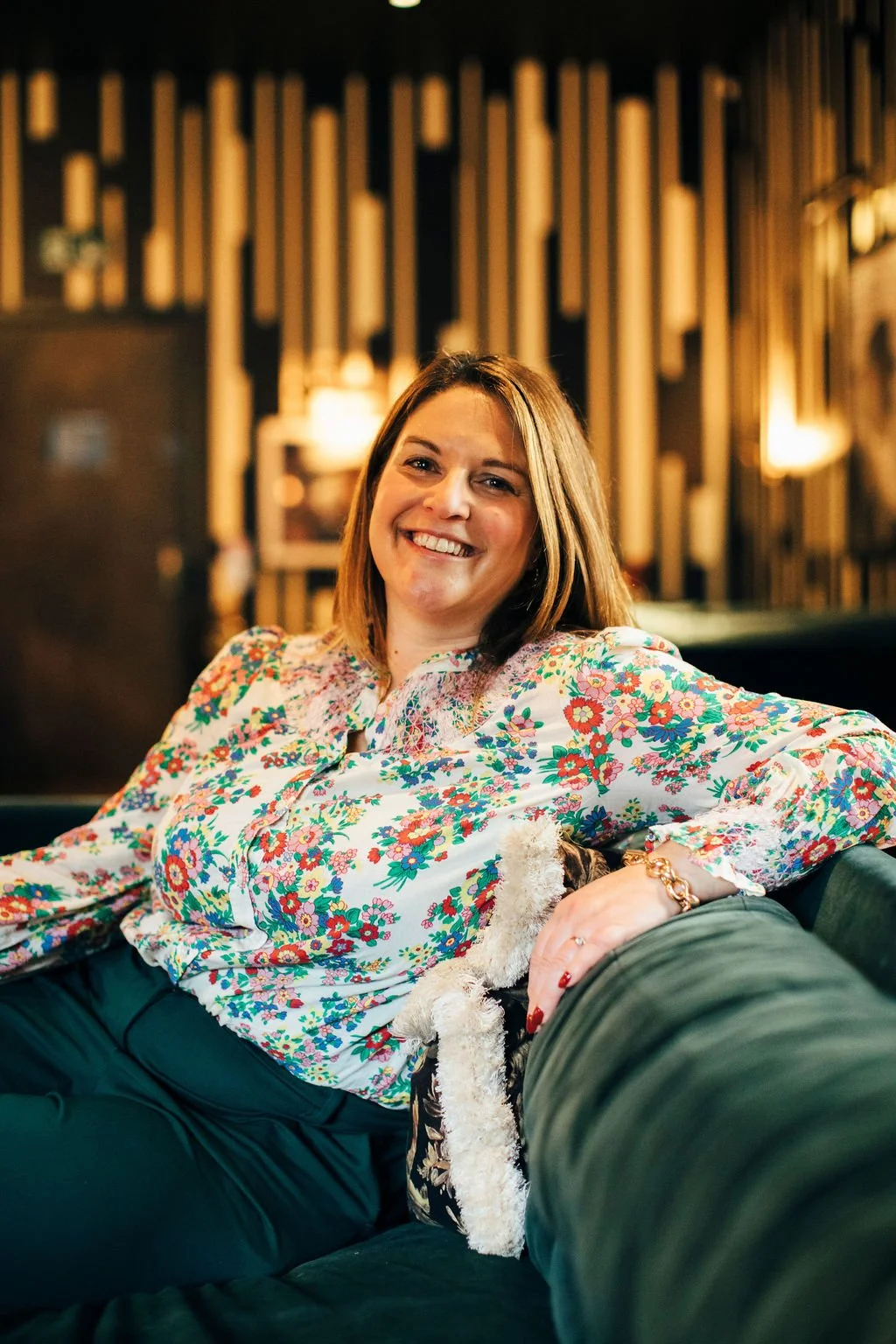 Becky Harrison Interior Designer sat on a green velvet sofa smiling looking directly at the camera. Wearing a flowery shirt, green trousers a chucky gold link bracelet and with red painted nails
