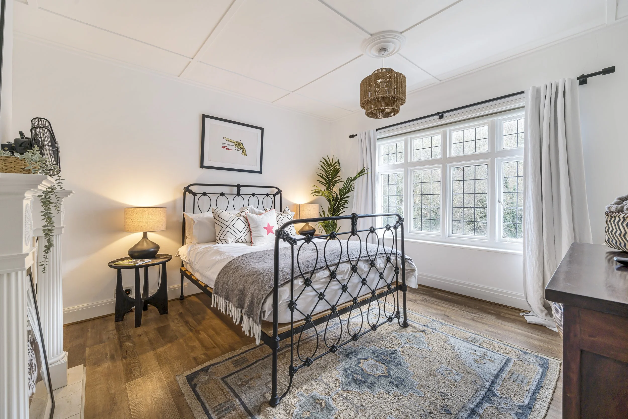 A large bedroom with white painted walls and a dark wooden floor with a vintage style faded rug. The room has a wrought iron double bed, 2 black mid century bedside tables with lamps & plants