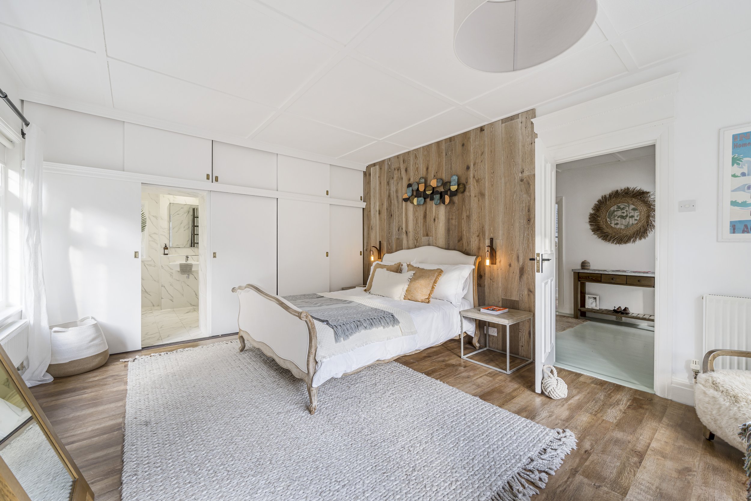 A large light filled bedrooom painted white. There are 2 open doors one to the hallway and one to the ensuite. There is a large king size bed. The room has a feature wall behind the bed where floorboards have been taken up the wall