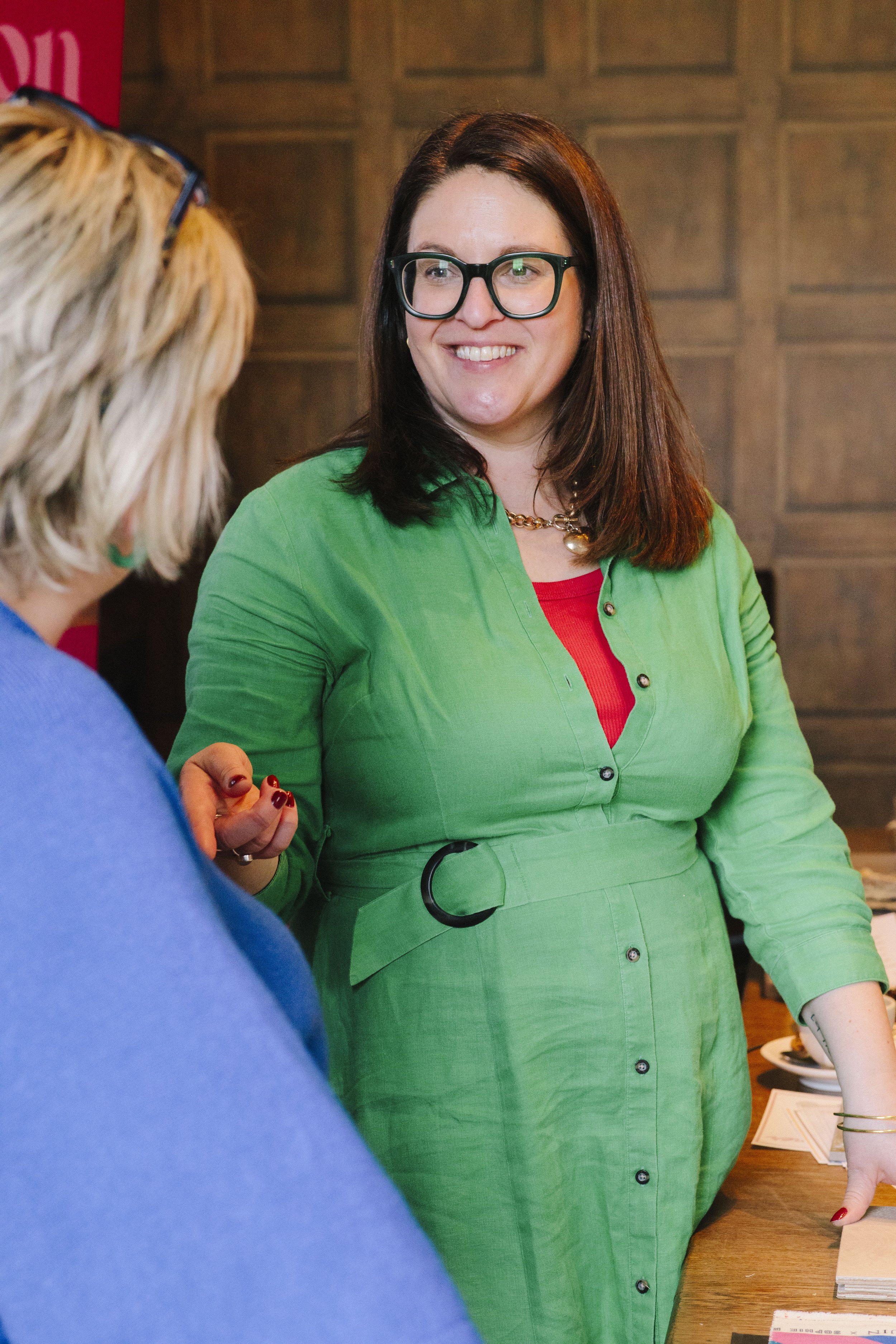 A smiling white woman wearing a green dress, red vest and green glasses speaks to another lady who has her back to the camera but is listening intently to the design advice being offered