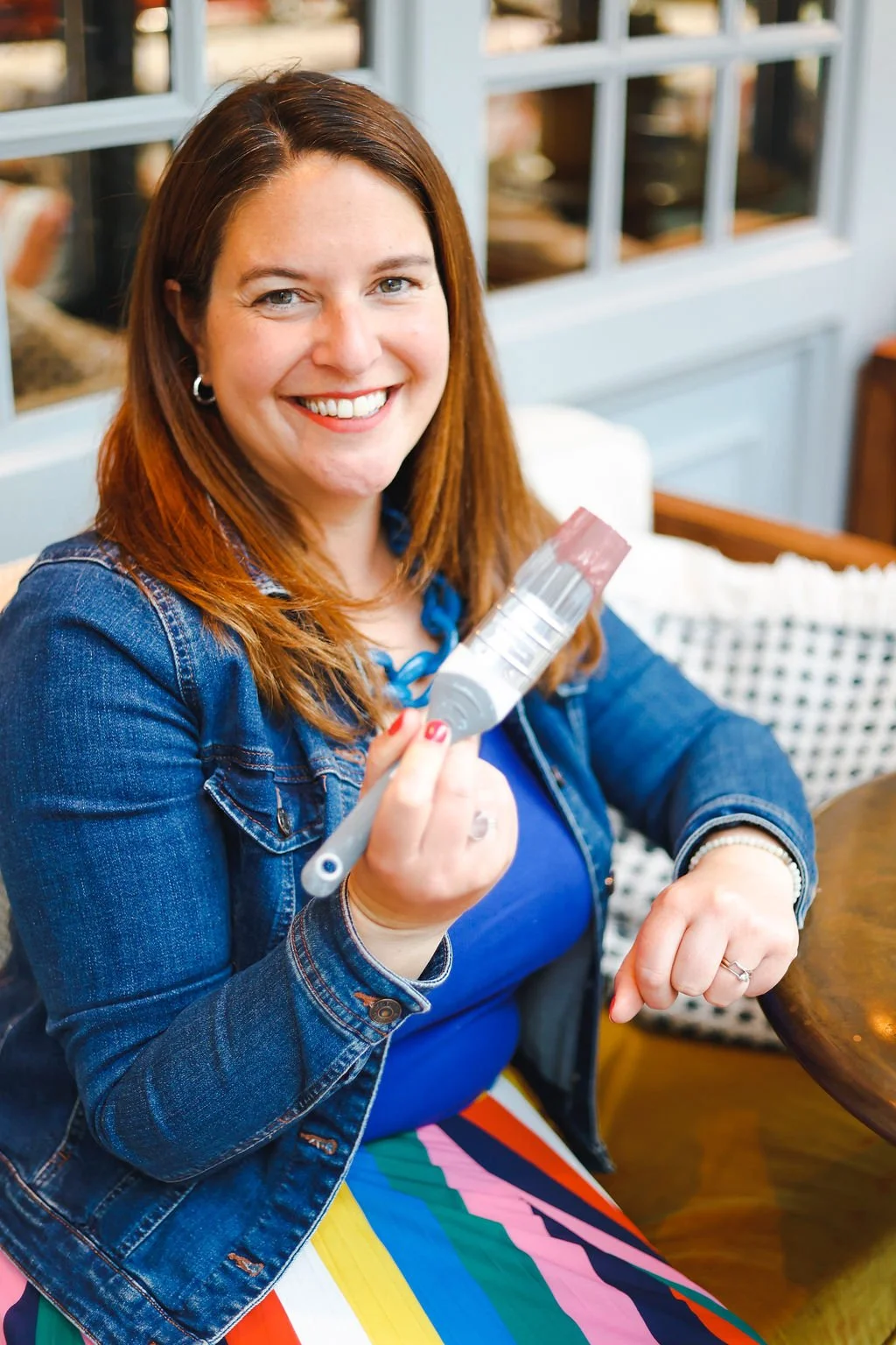 A female interior designer with brown hair and a large smile is sat at a dark wood table holdhing a paint brush wearing a multi coloured skirt