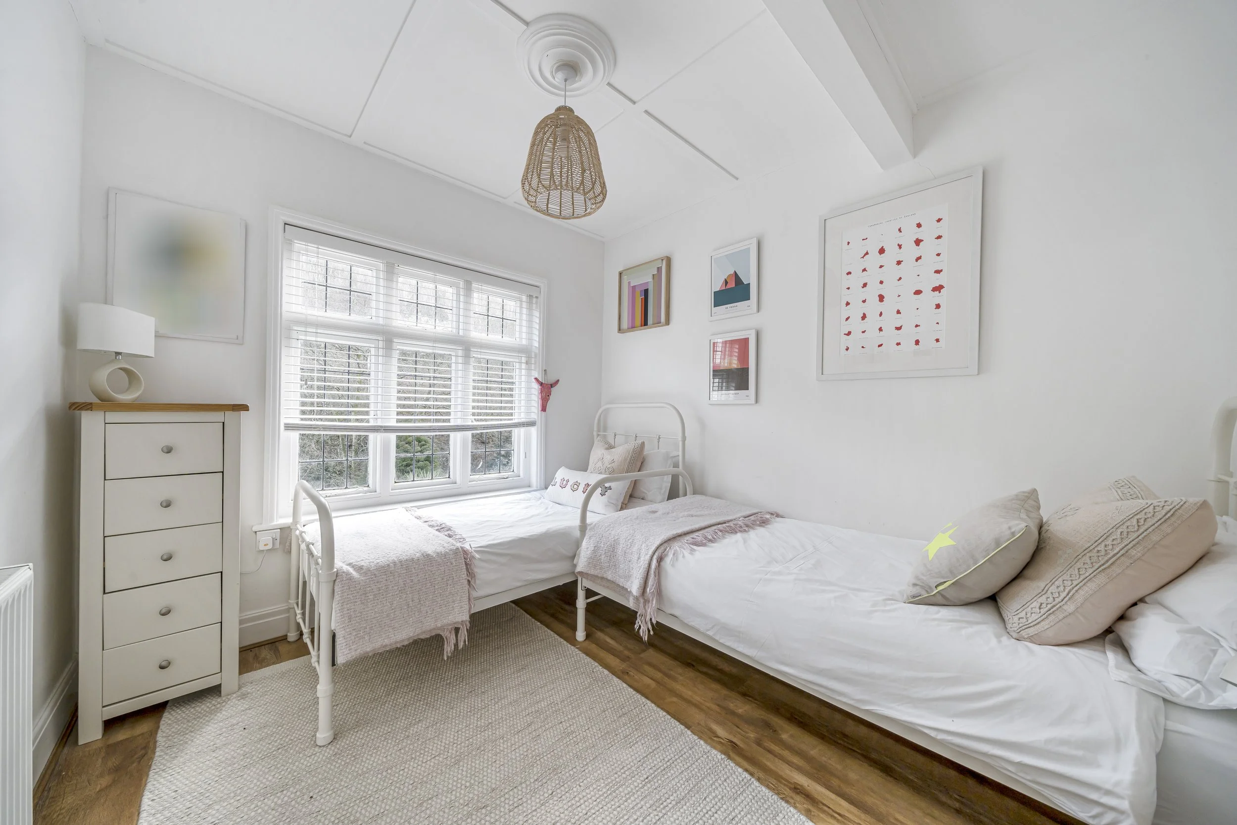 A bedroom with 2 single beds, the walls are painted white and the floor is wooden with a neutral tetured rug. The window is large and has a blind that is 2/3 closed. There is abstract wall art in bright pink & green colours.