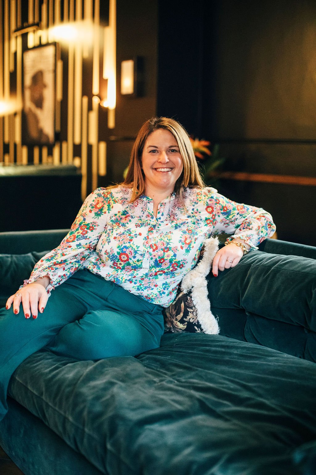 Becky Harrison Interior Designer sat on a green velvet sofa in a fairly dark room wearing a flowery short and green trousers