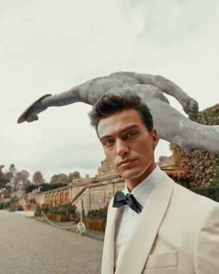 A man in a tuxedo standing outdoors with a large sculpture of a bird behind him.