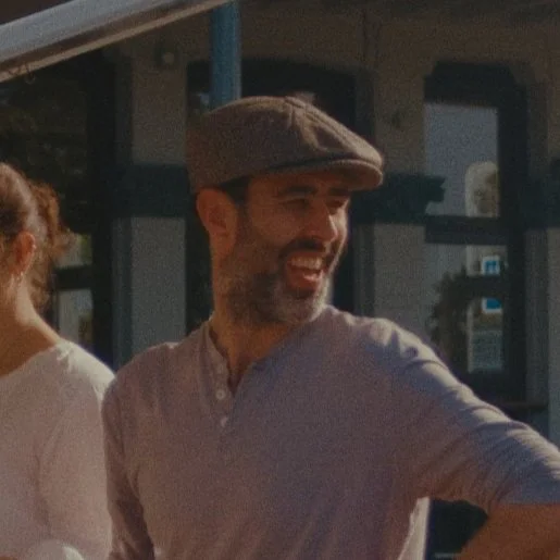 A man wearing a gray flat cap and light-colored shirt, smiling outdoors with a building in the background.