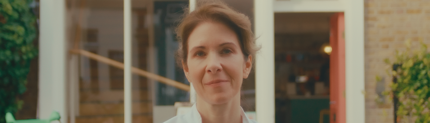 A woman with short brown hair standing outside near a house, smiling softly.